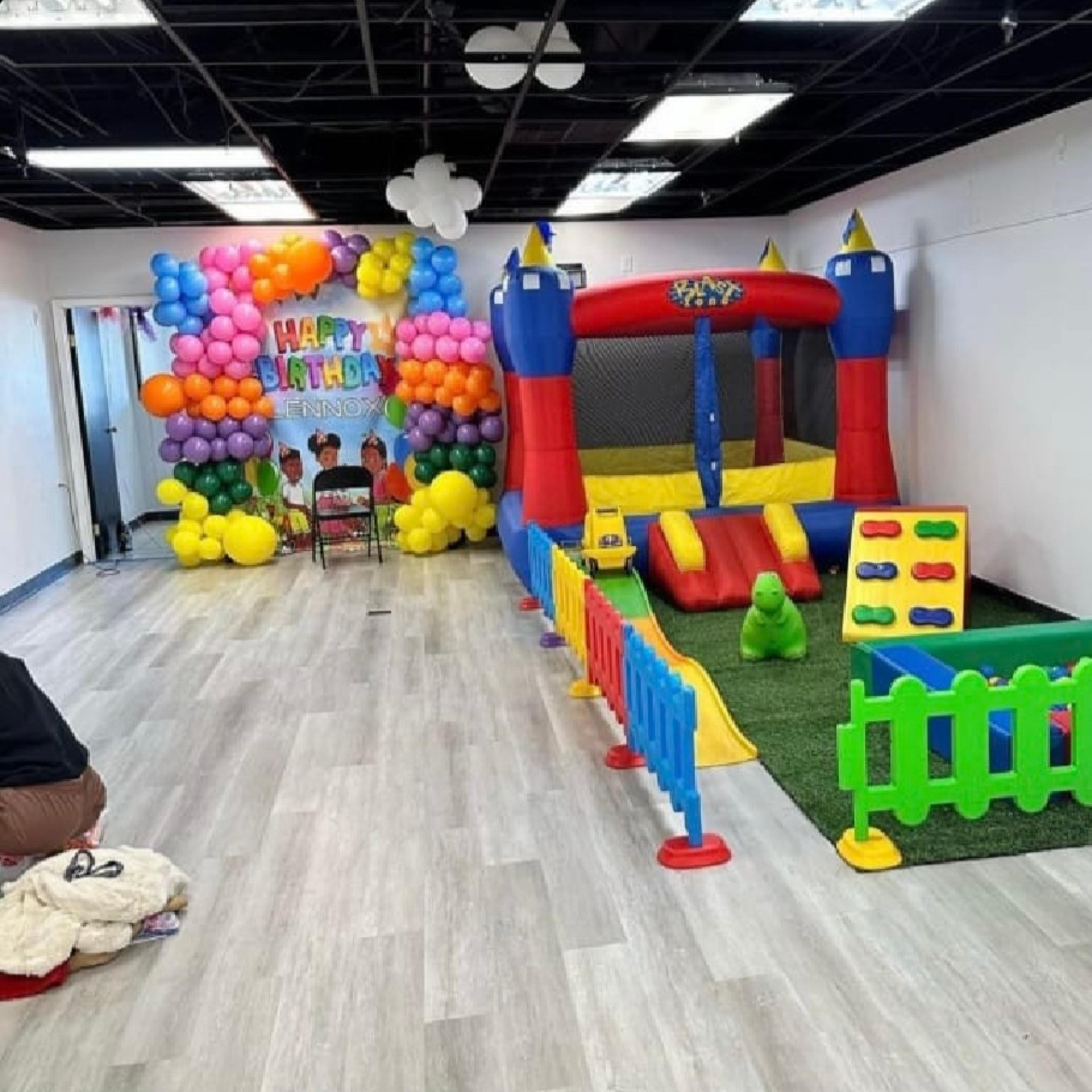 A colorful indoor party area with a bouncy castle, a play area surrounded by a toy fence, and a balloon backdrop that reads "Happy Birthday."