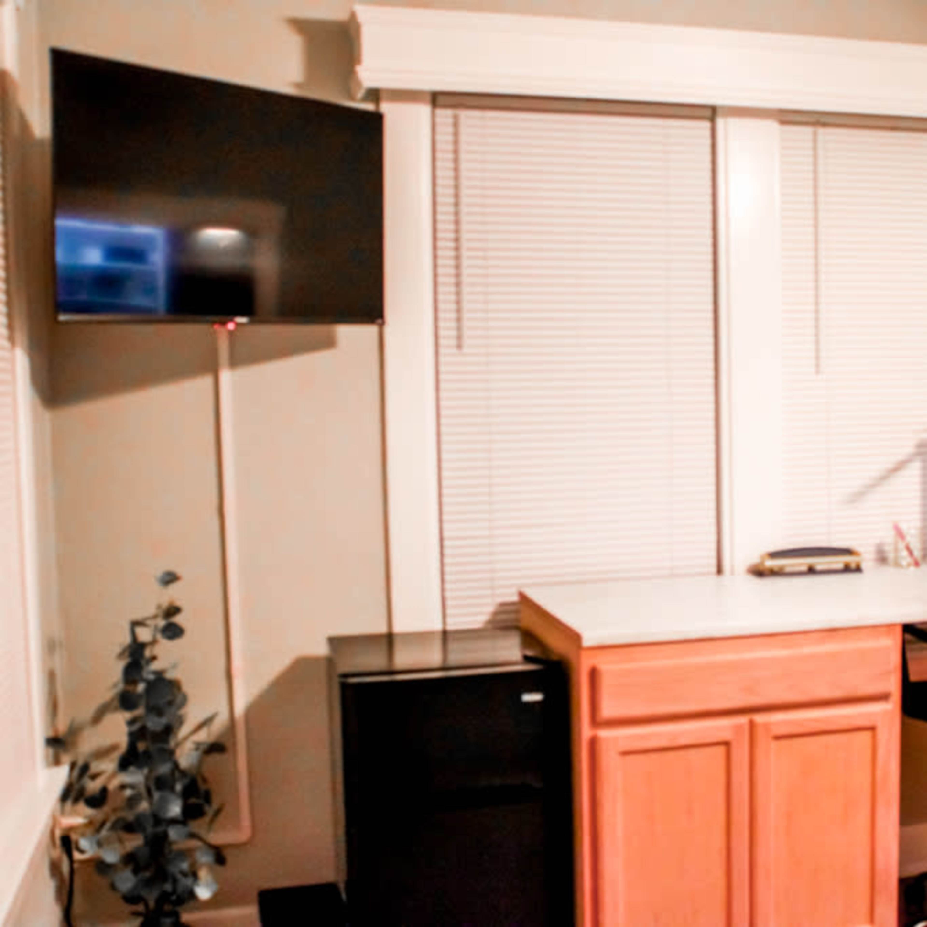 A home office with a desk, a black office chair, a small refrigerator, and a mounted flat-screen TV on the wall.