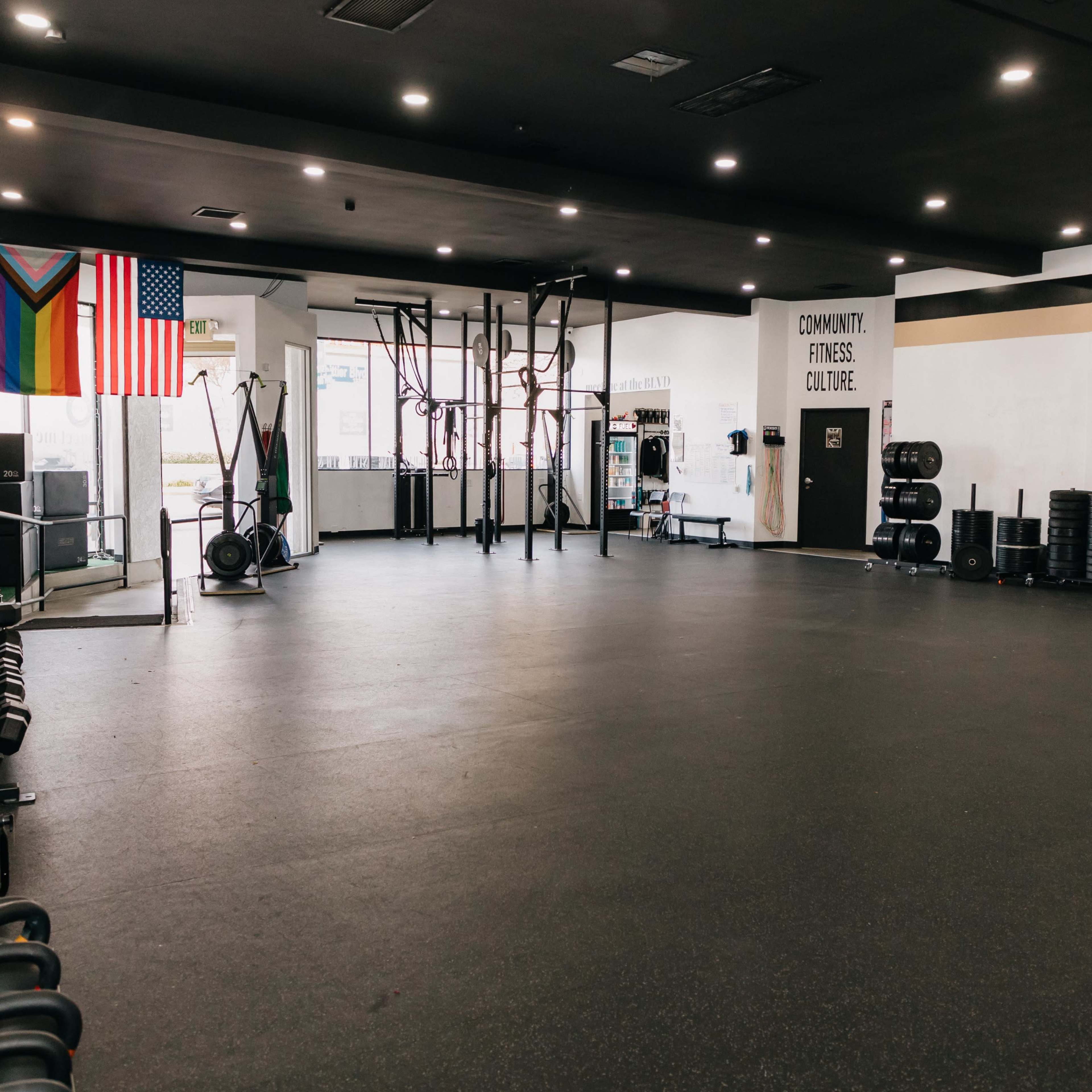 An empty gym with various exercise equipment, including dumbbells on racks, a set of pull-up bars, and wall-mounted storage, alongside flags hanging from the wall.