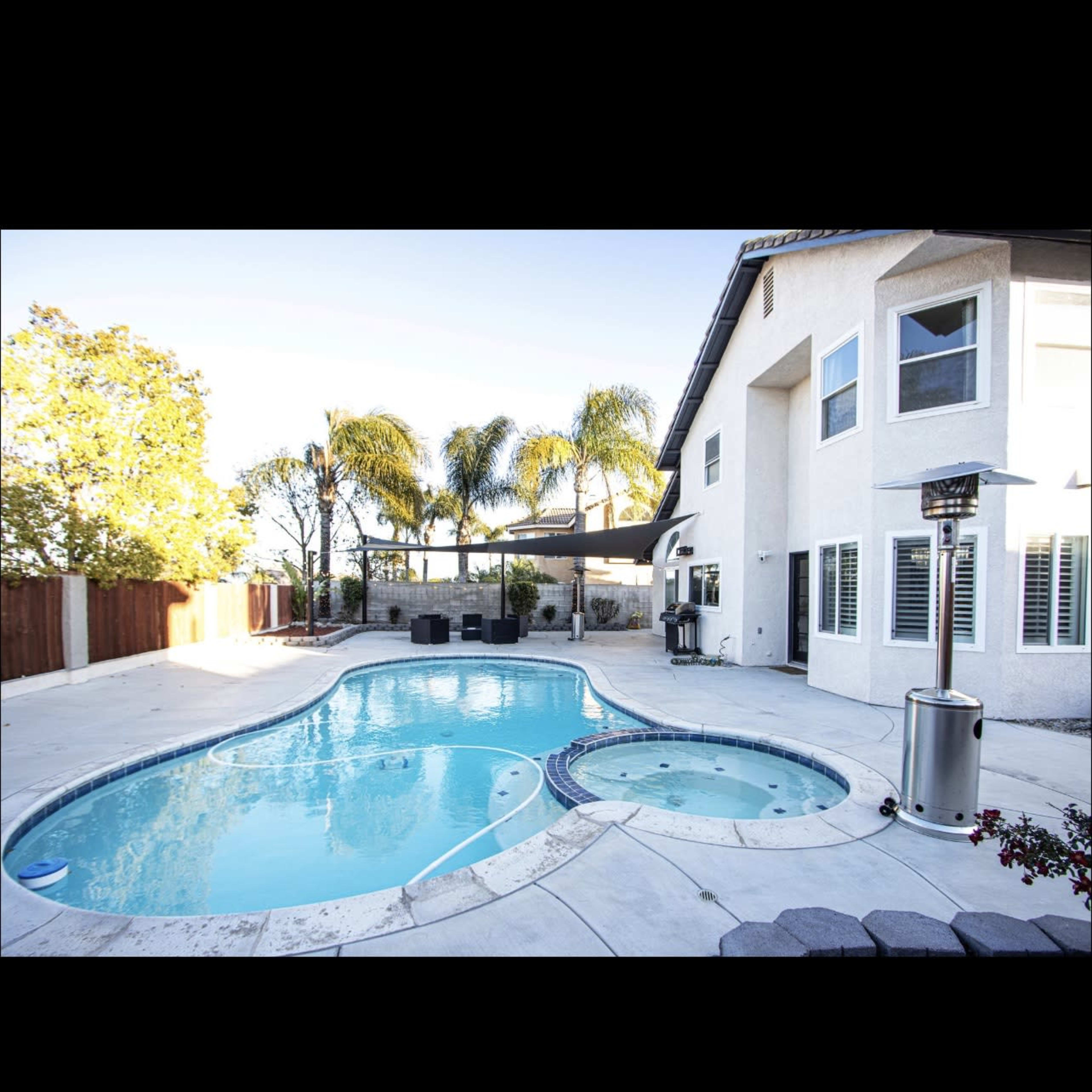 A backyard with a swimming pool and a spa, surrounded by palm trees and a white house.