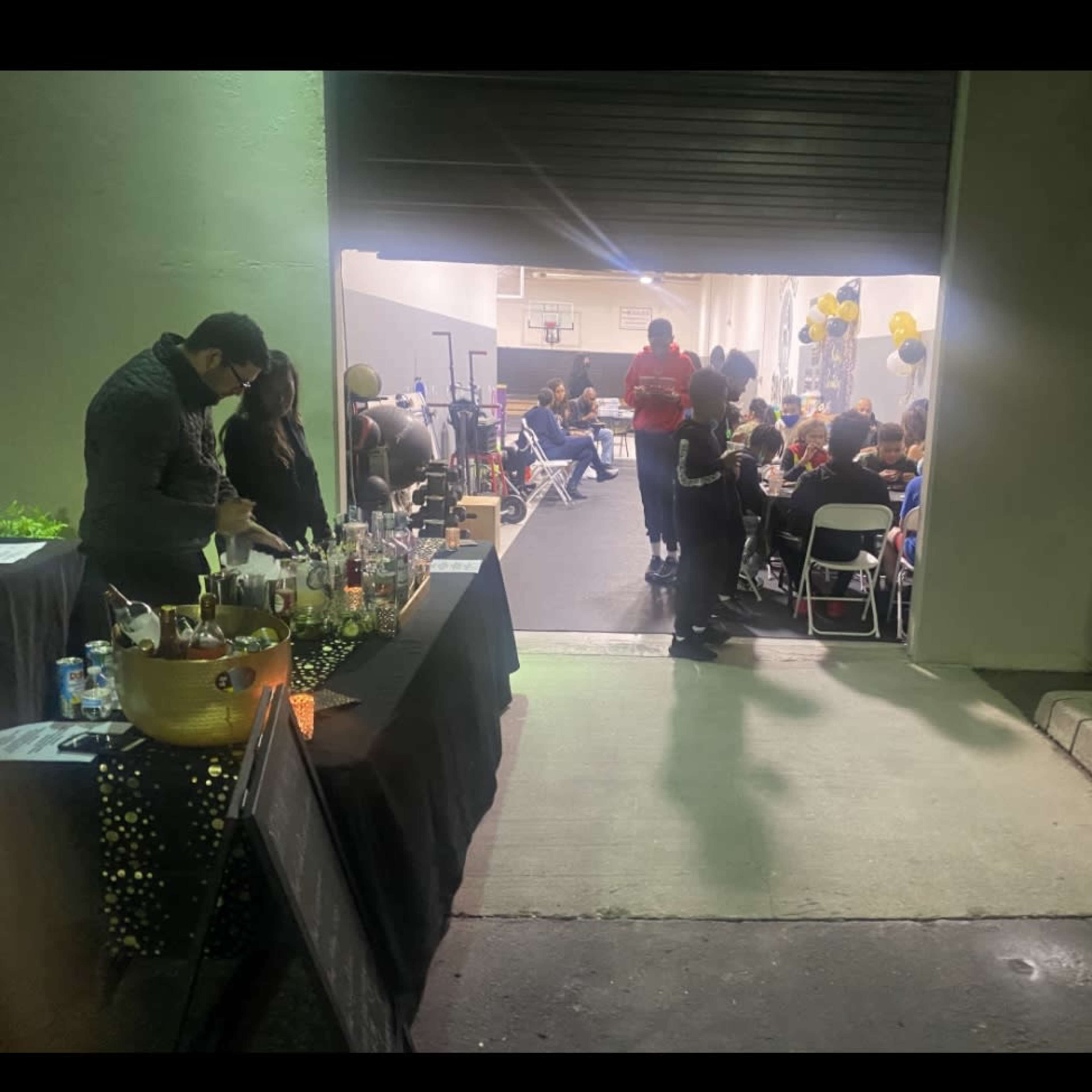 A group of people is gathered at a festive event inside a dimly lit venue, with a bartender serving drinks at a table set up outside the entrance.