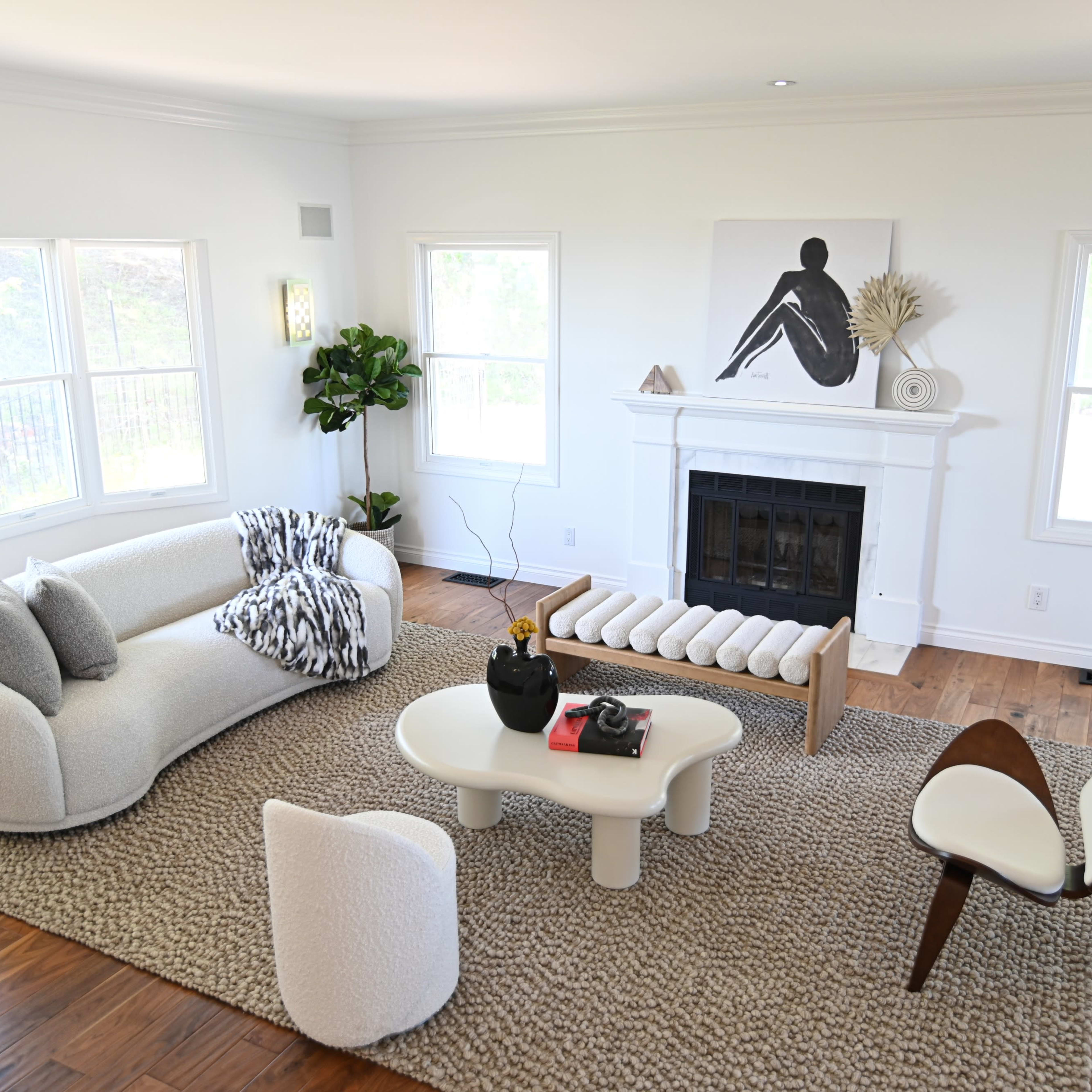 A modern living room with a curved sofa, a wooden coffee table, a patterned rug, and large windows offering natural light.