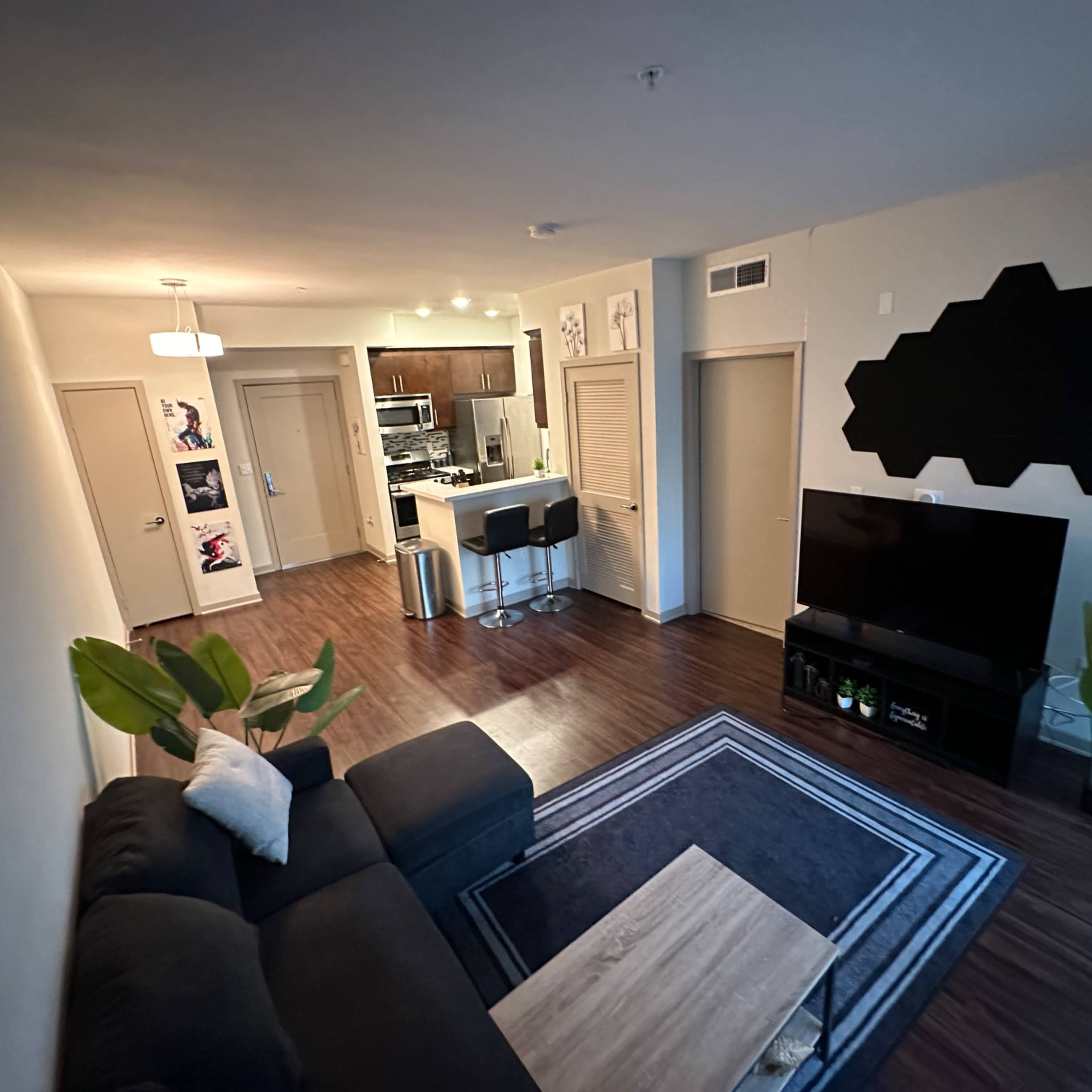 A modern living room with a dark gray sectional sofa, a coffee table, and a TV stand, leading into an open kitchen area with bar stools.