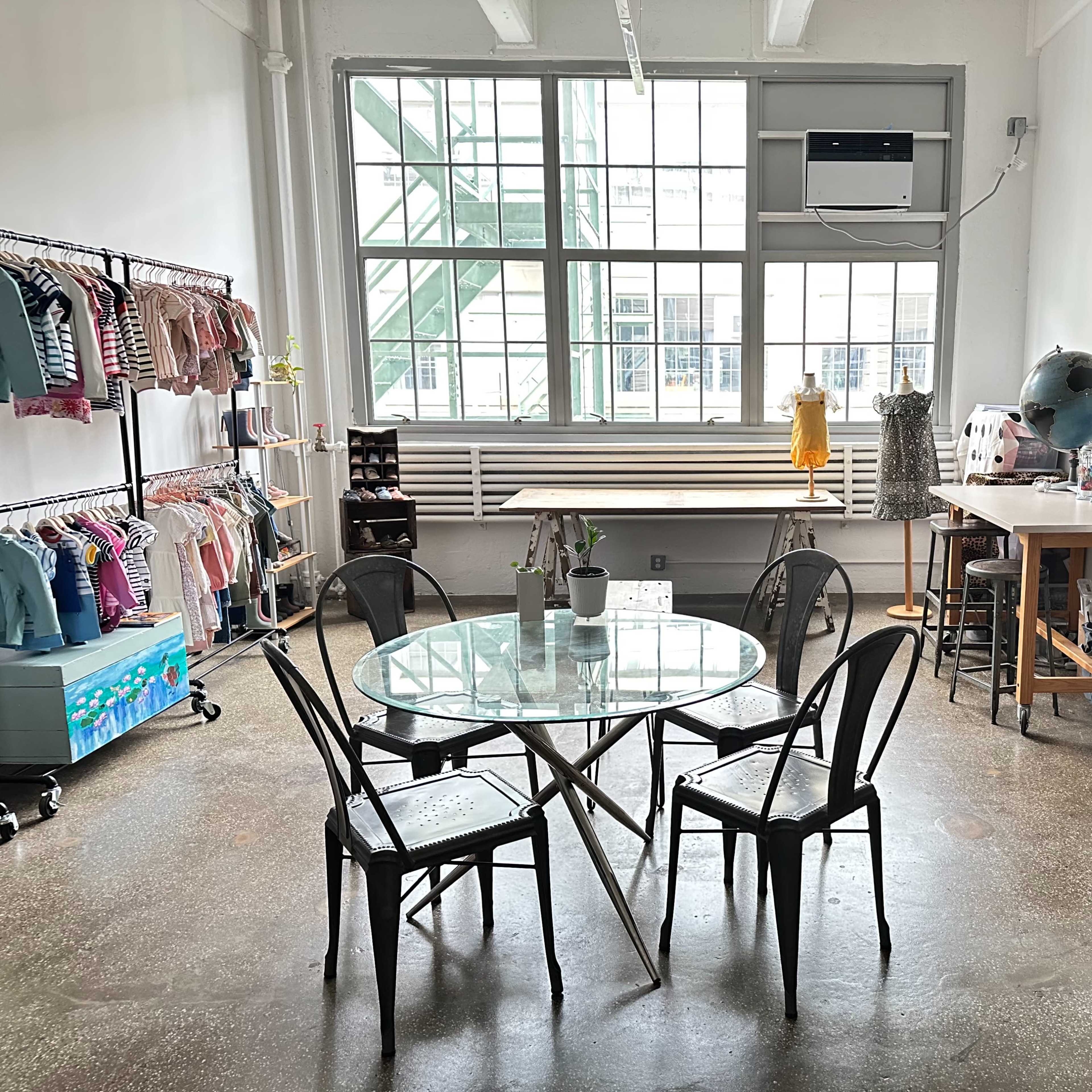 A bright clothing store interior with a circular glass table surrounded by black chairs, clothing racks on the walls, and a worktable in the corner.