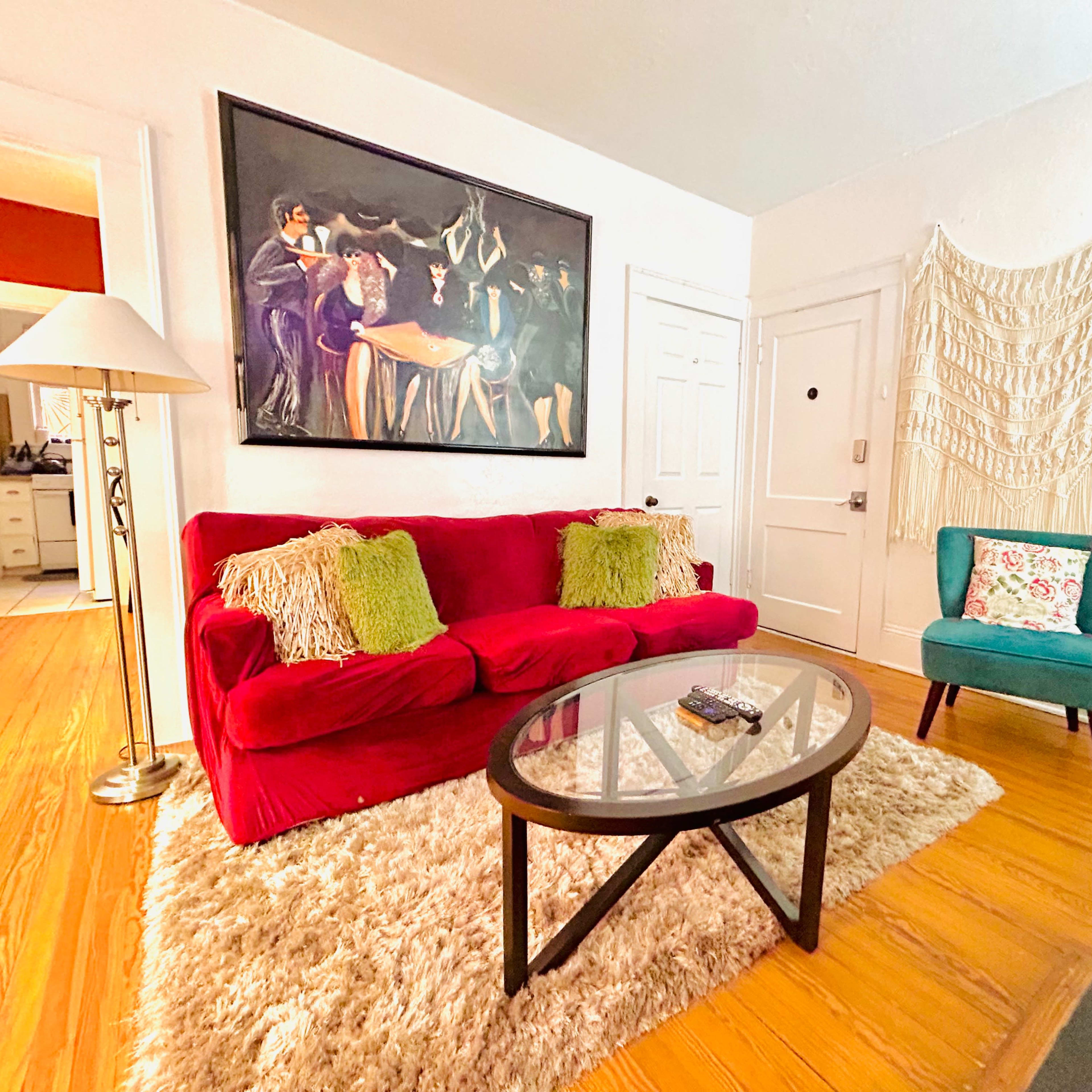 A living room features a red sofa with decorative pillows, a glass coffee table, and a large painting, alongside a teal chair and a textured wall hanging.