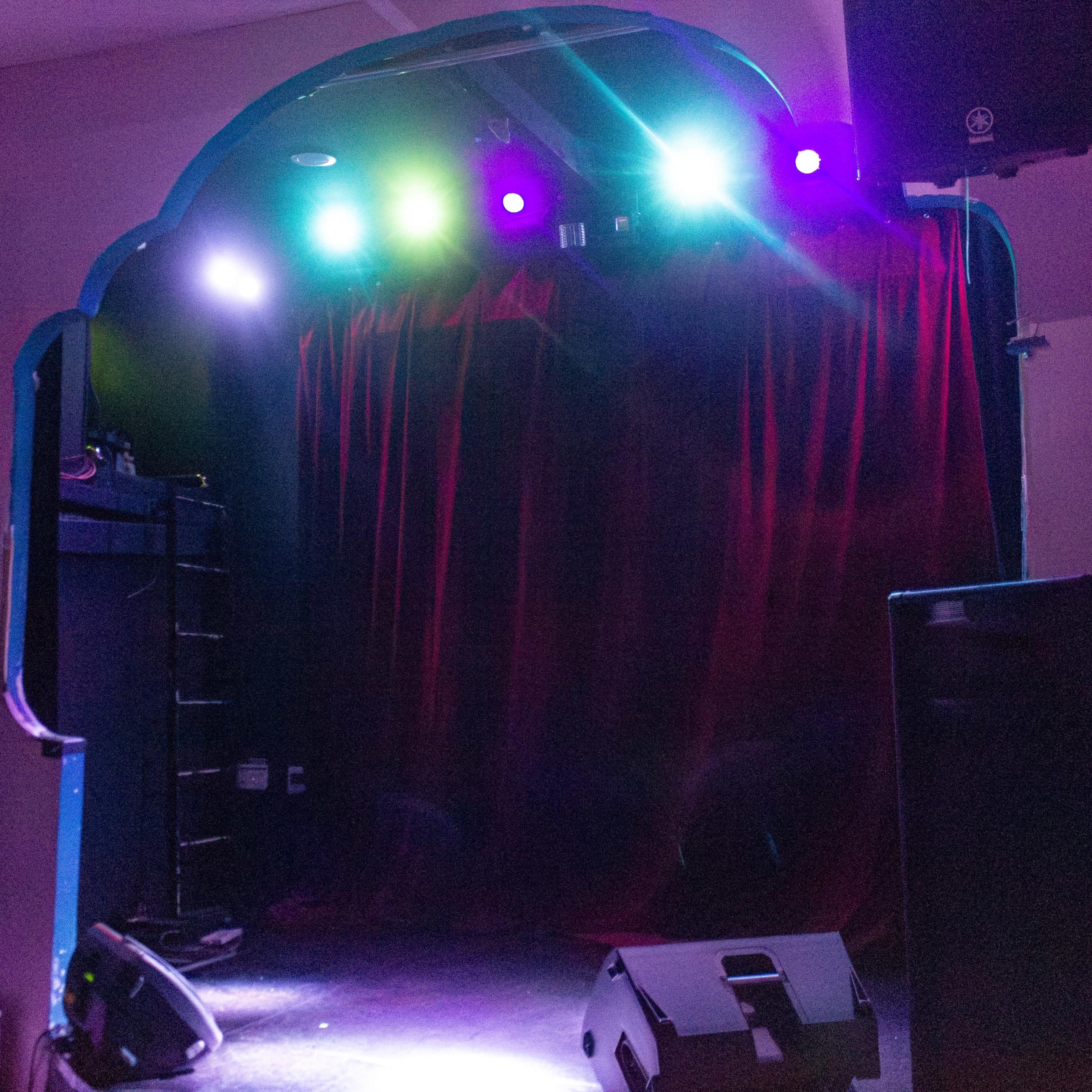 A stage with a red curtain, flanked by large speakers, and illuminated by colorful lights.