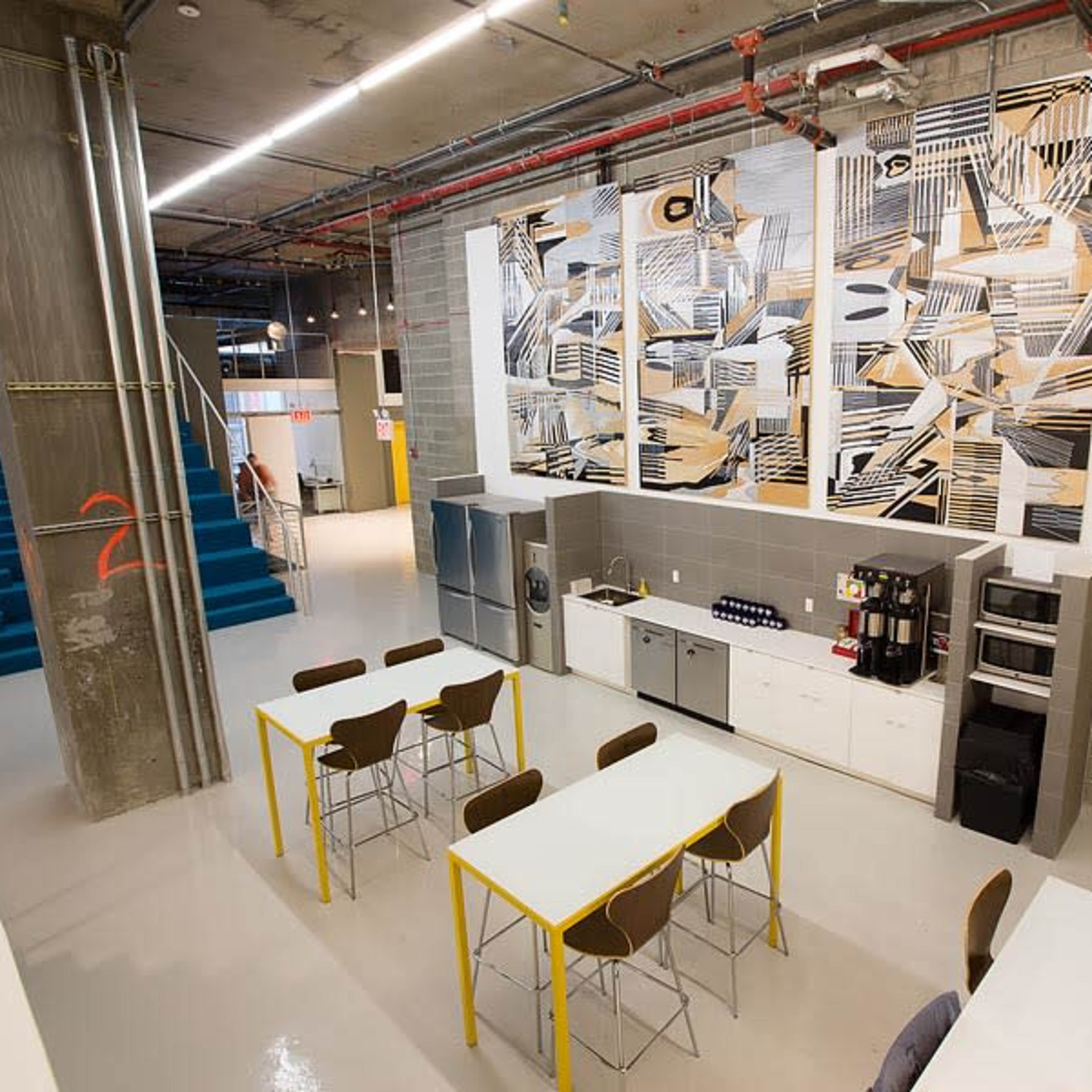A modern communal kitchen area with a large mural, several tables with chairs, and kitchen appliances along a wall.