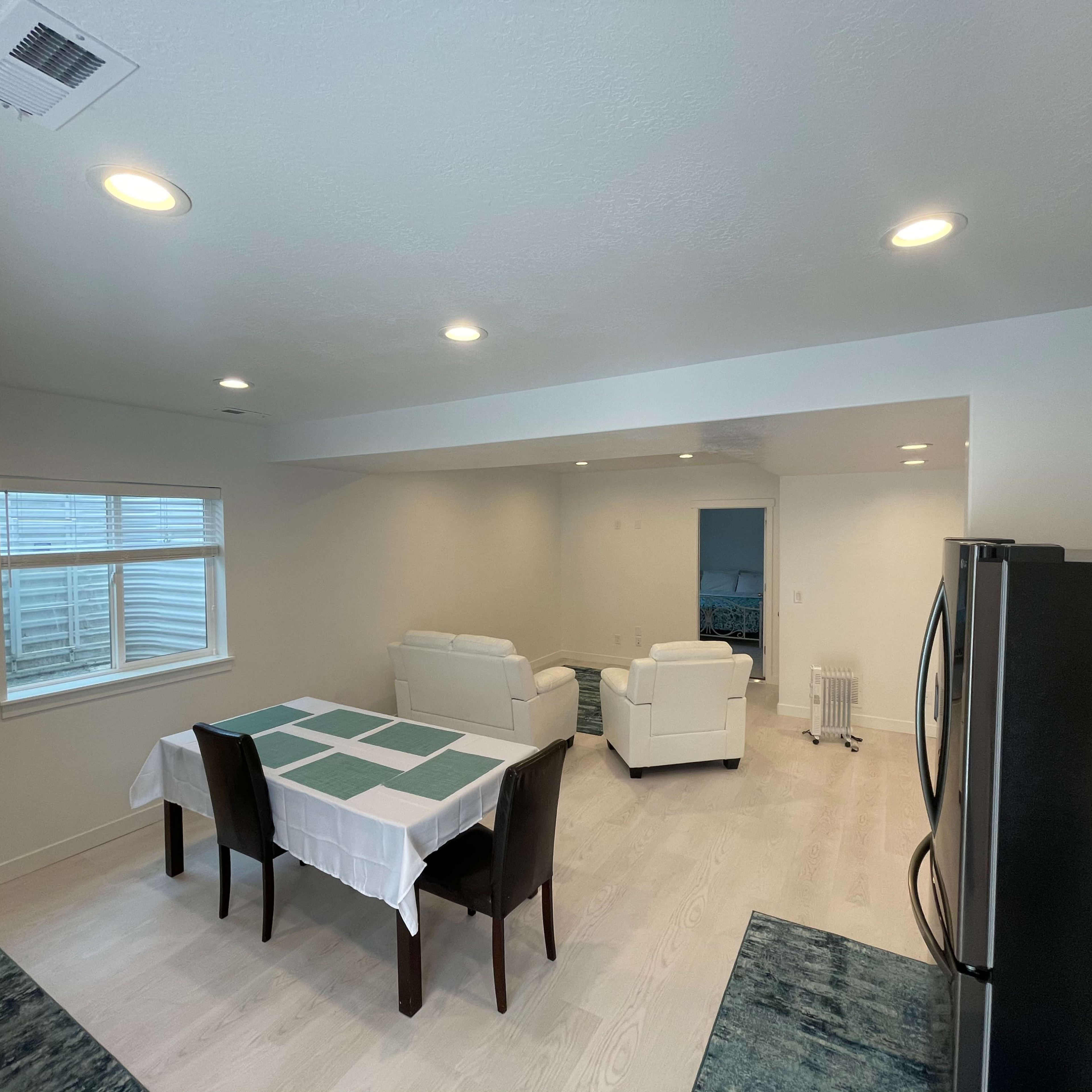 A modern kitchen and dining area featuring a table with a white tablecloth, four chairs, and two sofas in a well-lit, open-layout space.