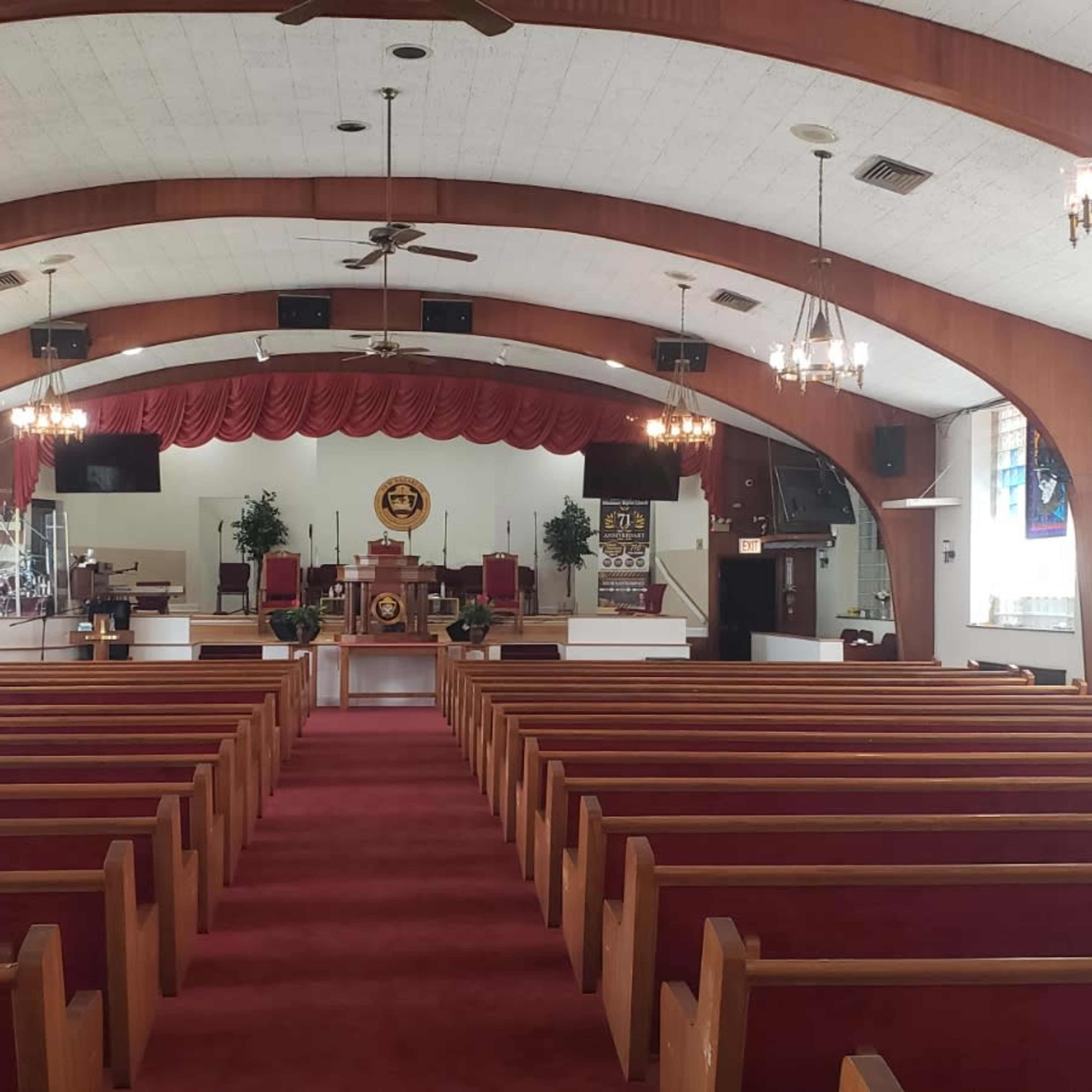 The interior of a church features rows of wooden pews facing a raised altar area with a lectern and decorative elements, under a vaulted ceiling.