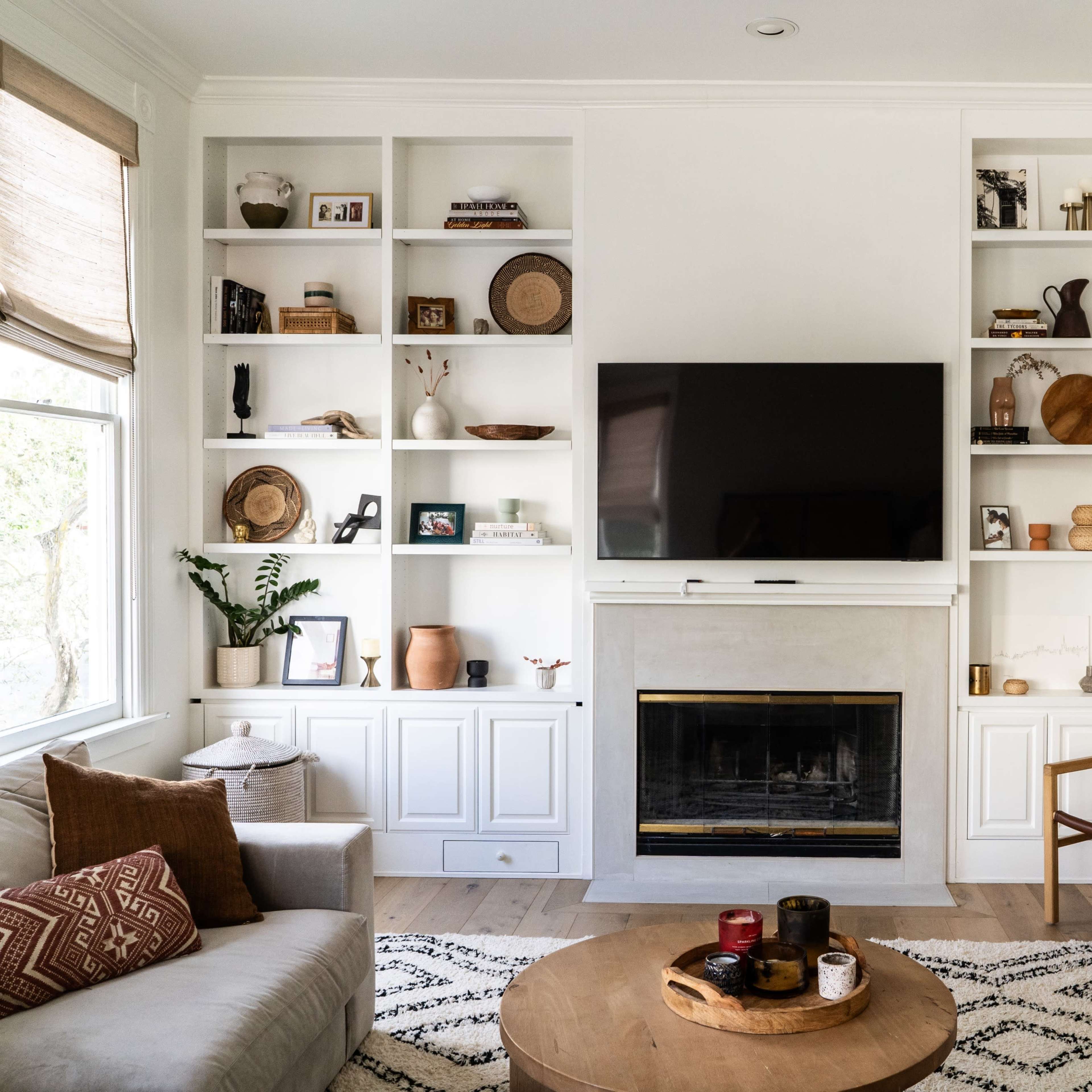 A living room features built-in shelves filled with decorative items, a television mounted above a fireplace, and a coffee table in front of a beige sofa.