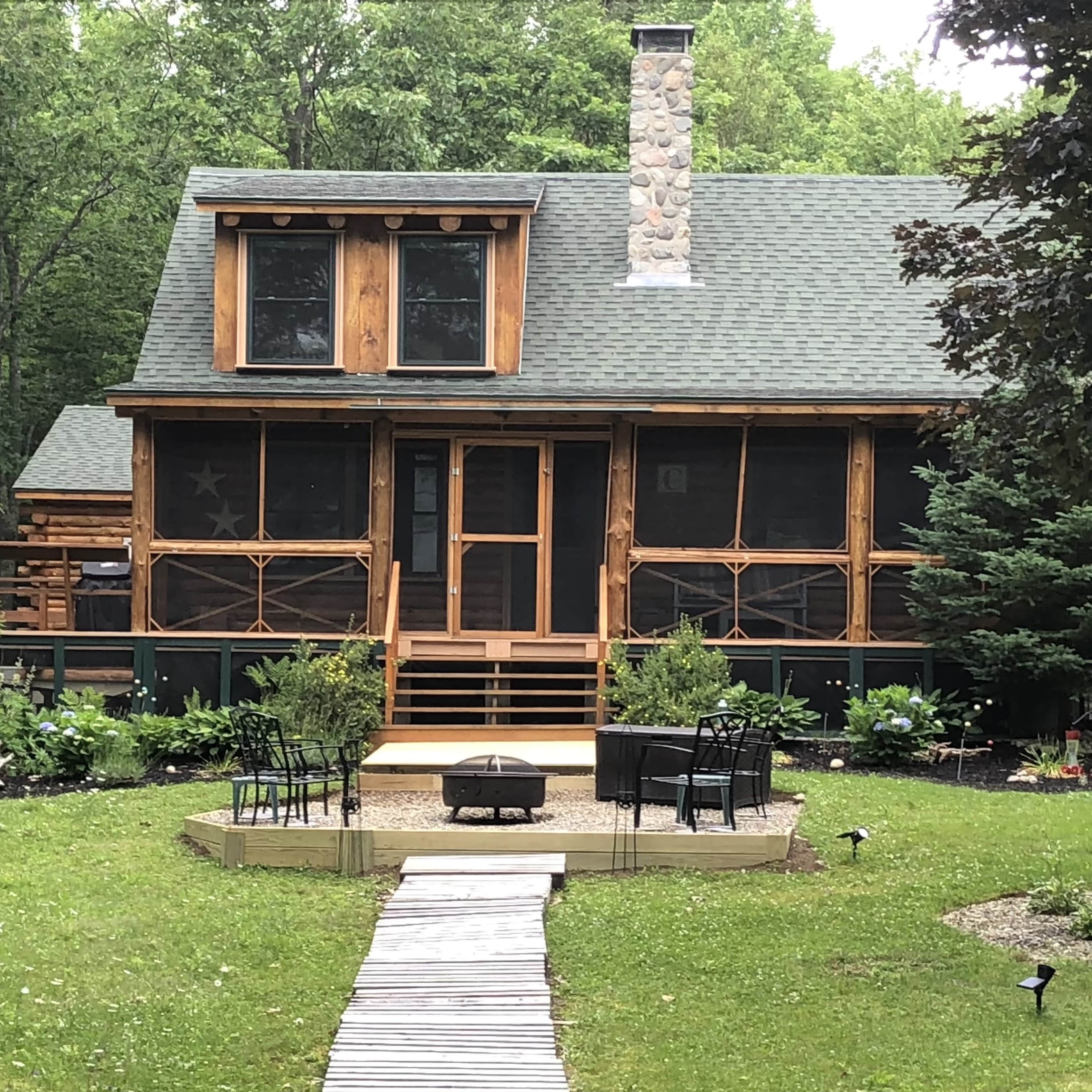 A rustic wooden cabin surrounded by greenery features a screened porch and a central outdoor fire pit with seating.