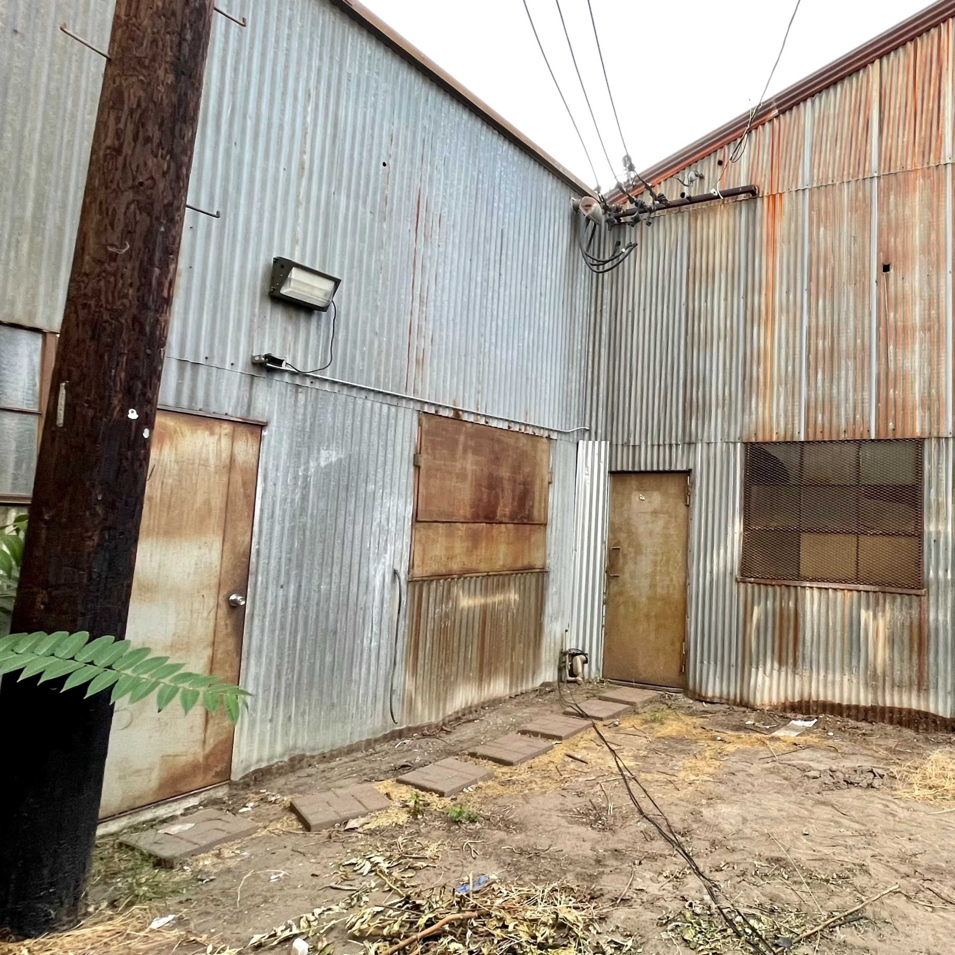 A narrow alley between two corrugated metal buildings, with a wooden post and overgrown weeds on the ground.