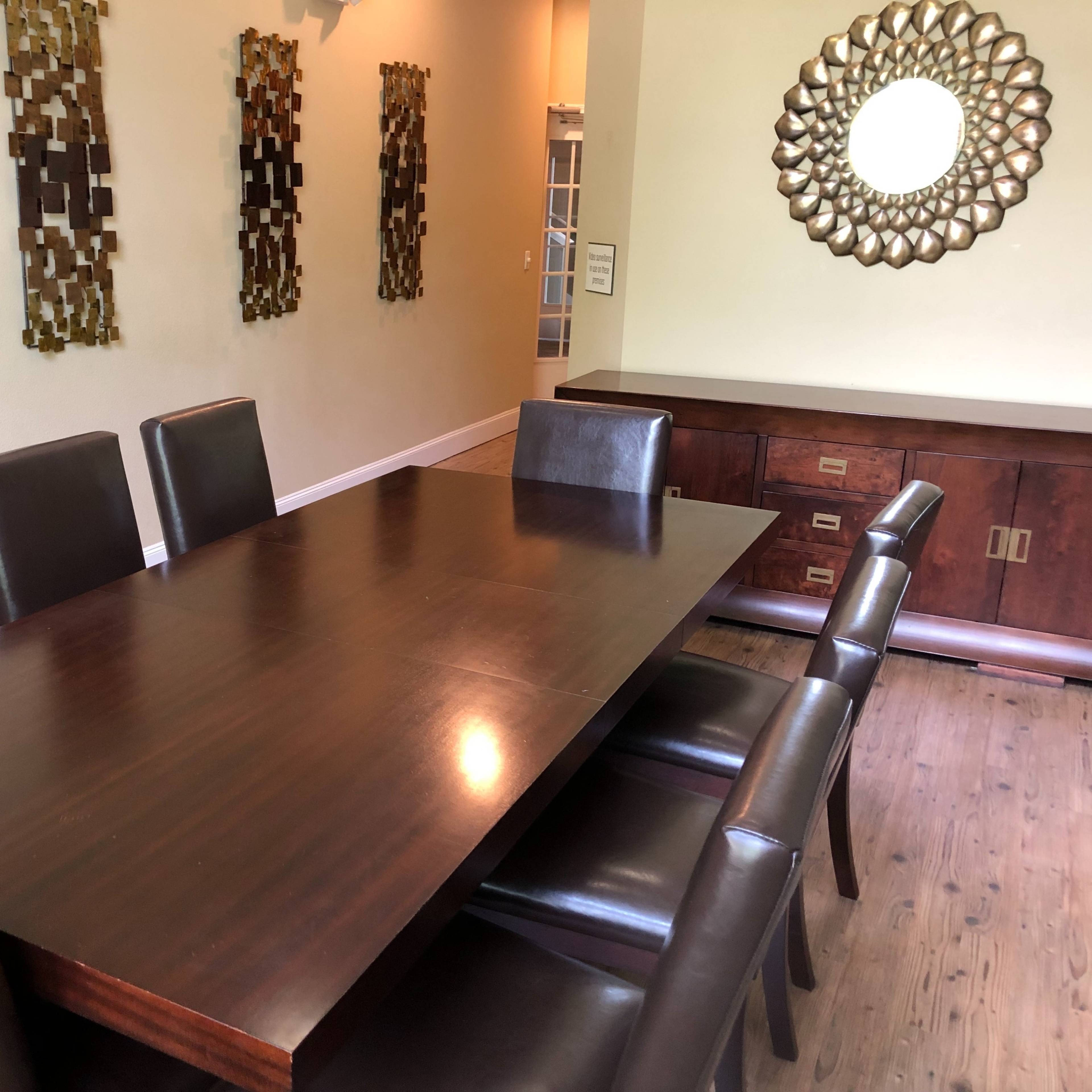 A rectangular wooden dining table with eight leather chairs is shown in a room featuring textured wall art and a circular mirror.