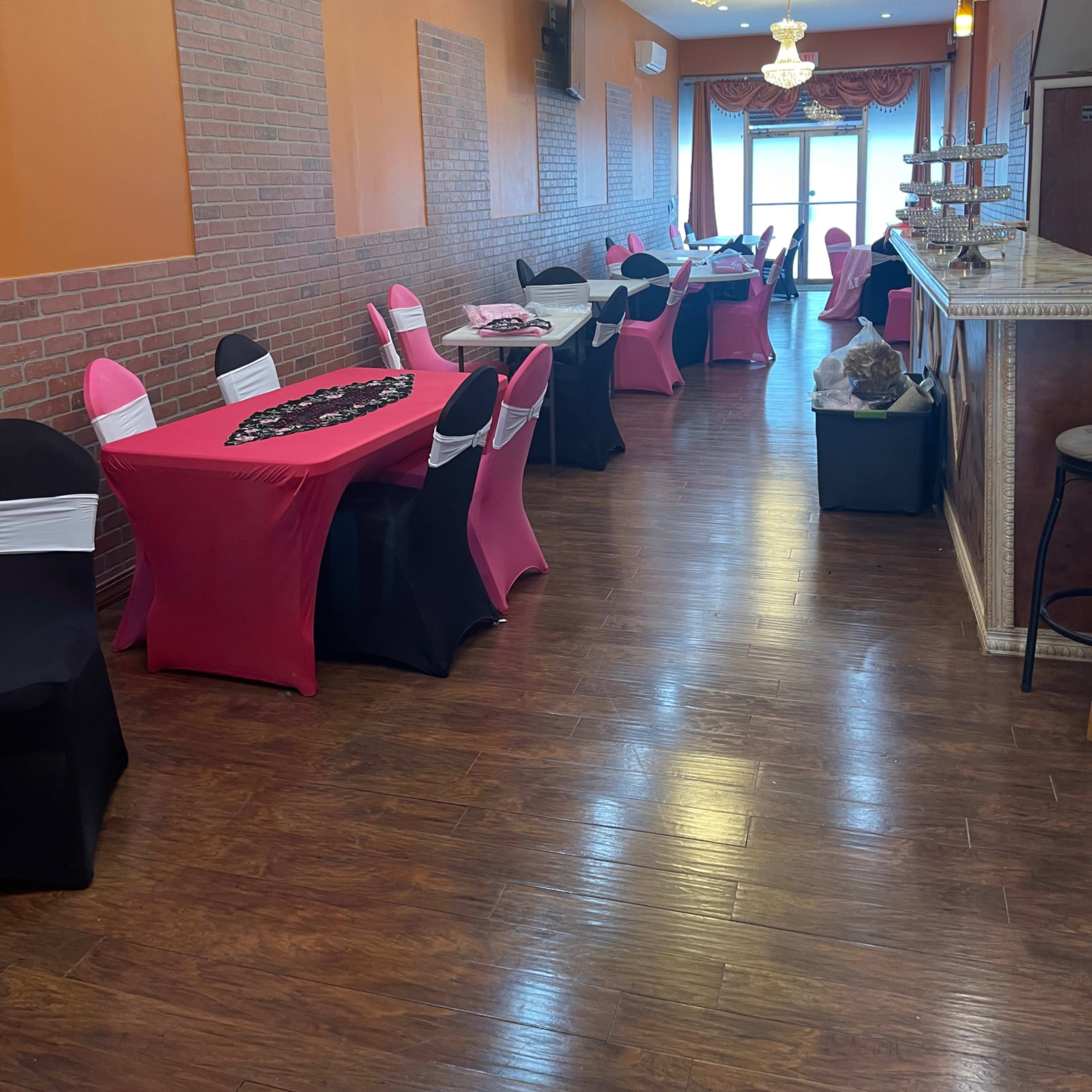 The interior of a restaurant features rows of tables dressed in black and pink tablecloths, with decorative lighting and a bar at the far end.