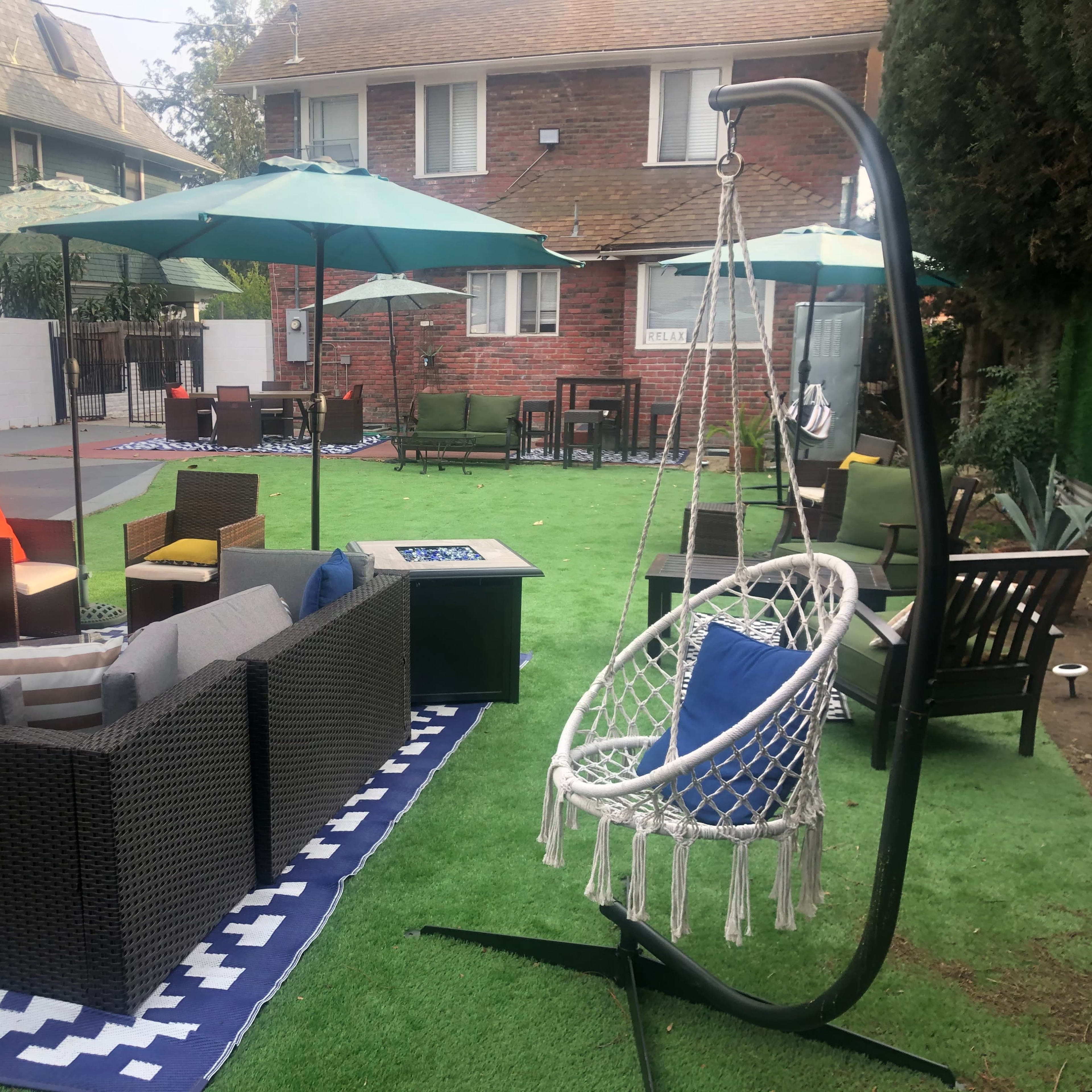 The scene features a landscaped outdoor area with seating arrangements, a swing chair, and umbrellas in a residential setting.