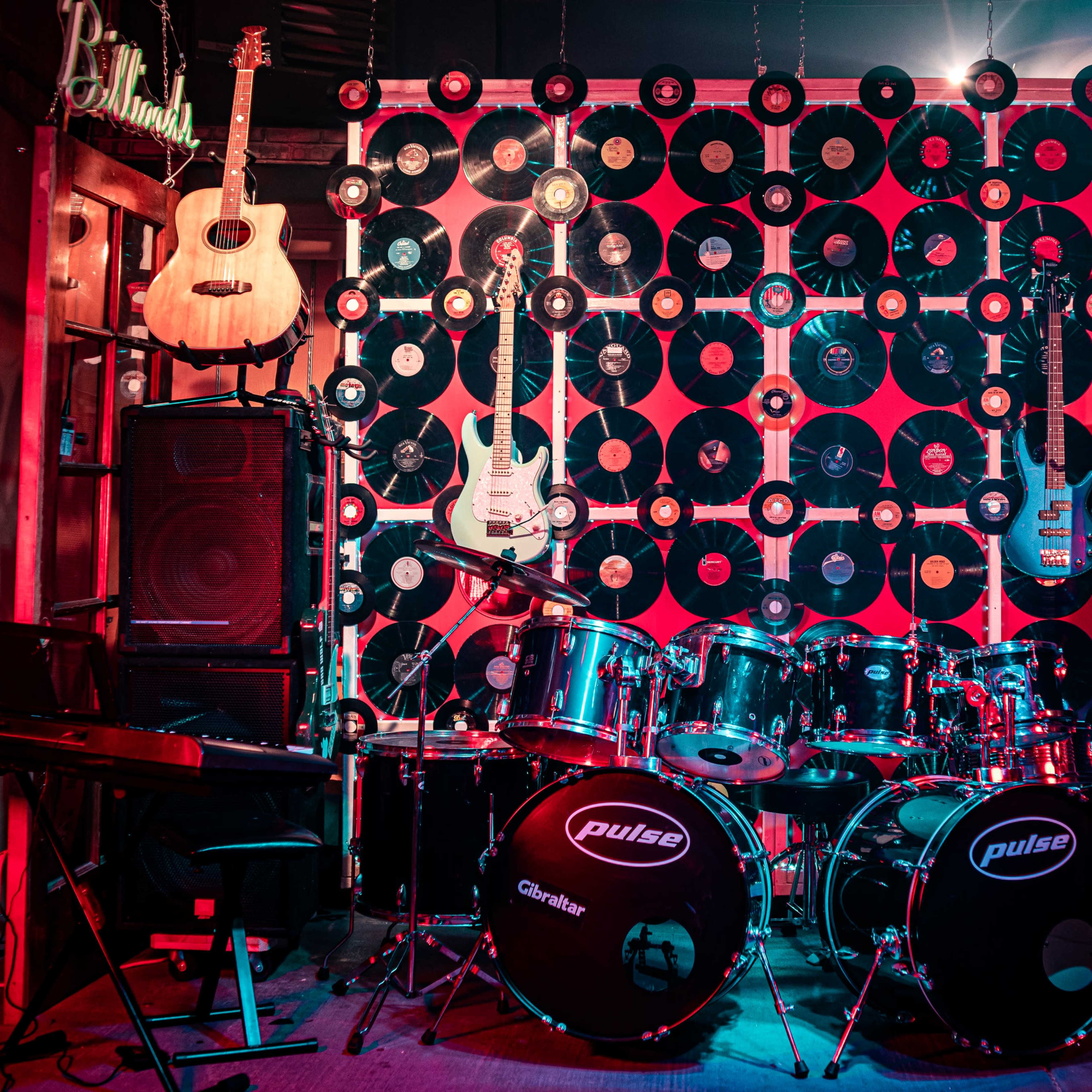The scene features a display of musical instruments, including guitars and a drum set, set against a backdrop of vinyl records and speakers.