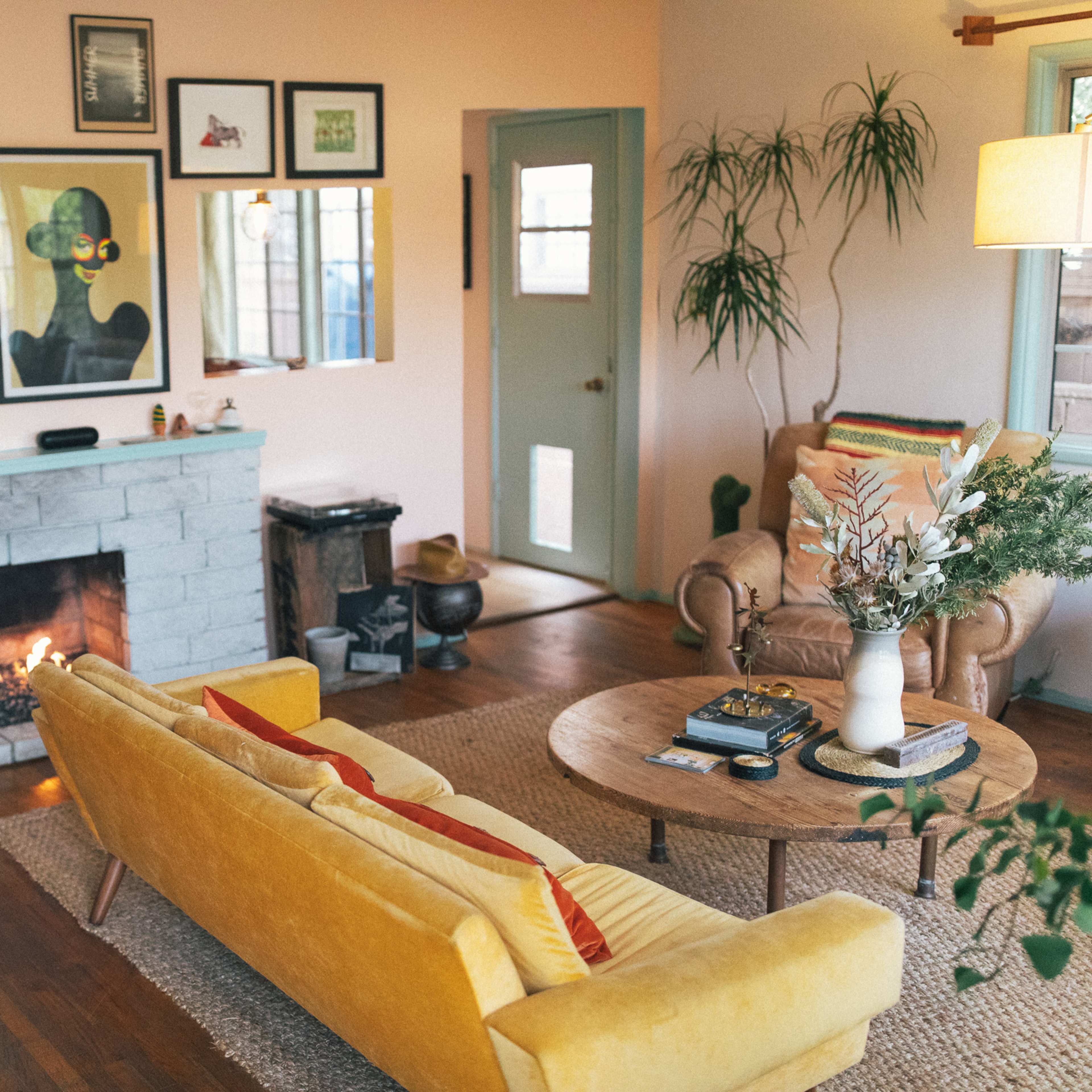 The scene features a cozy living room with a yellow couch, a wooden coffee table, an art-adorned wall, a fireplace, and various plants.