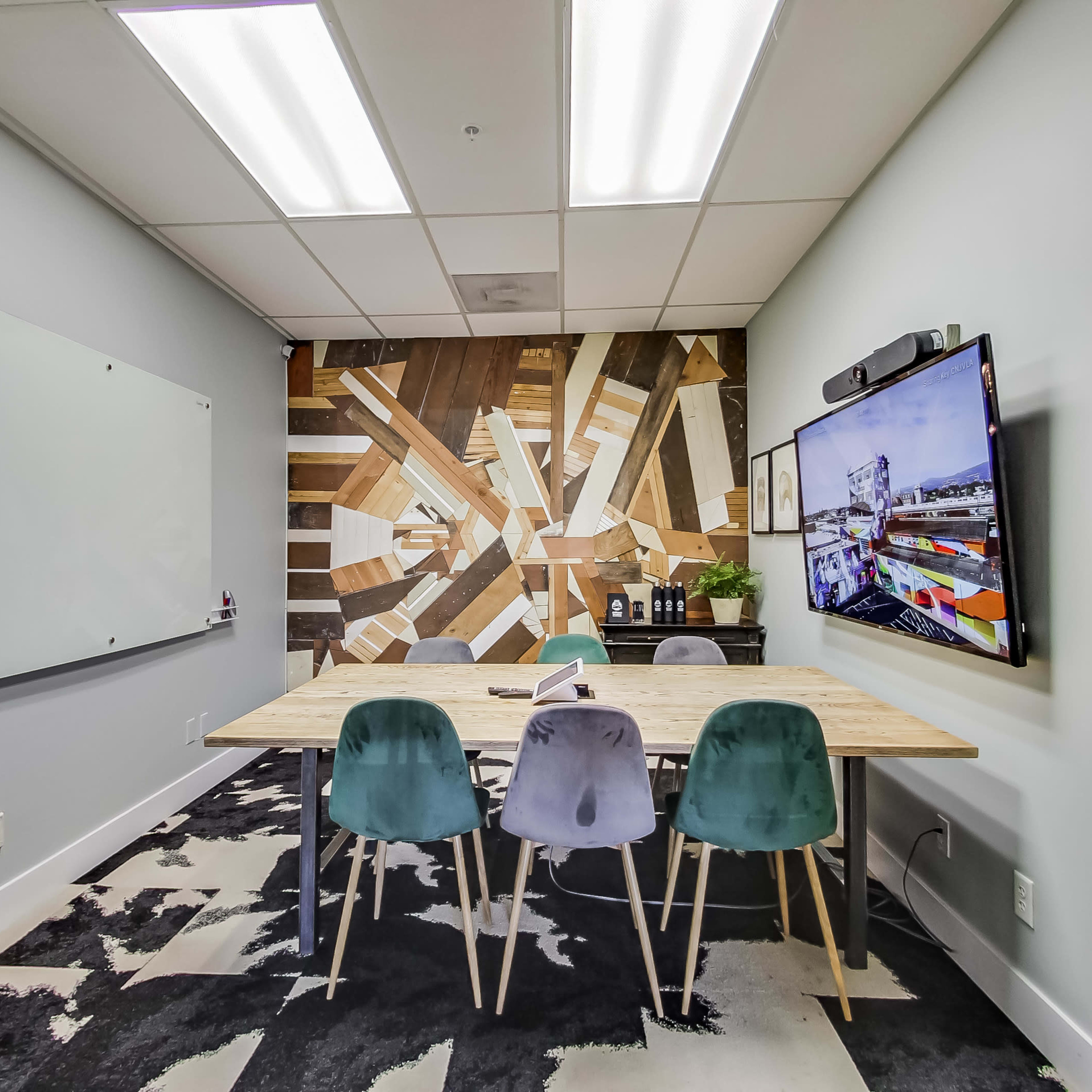 A modern conference room with a wooden table, four chairs, a large wall-mounted screen, and a decorative wood-patterned wall.