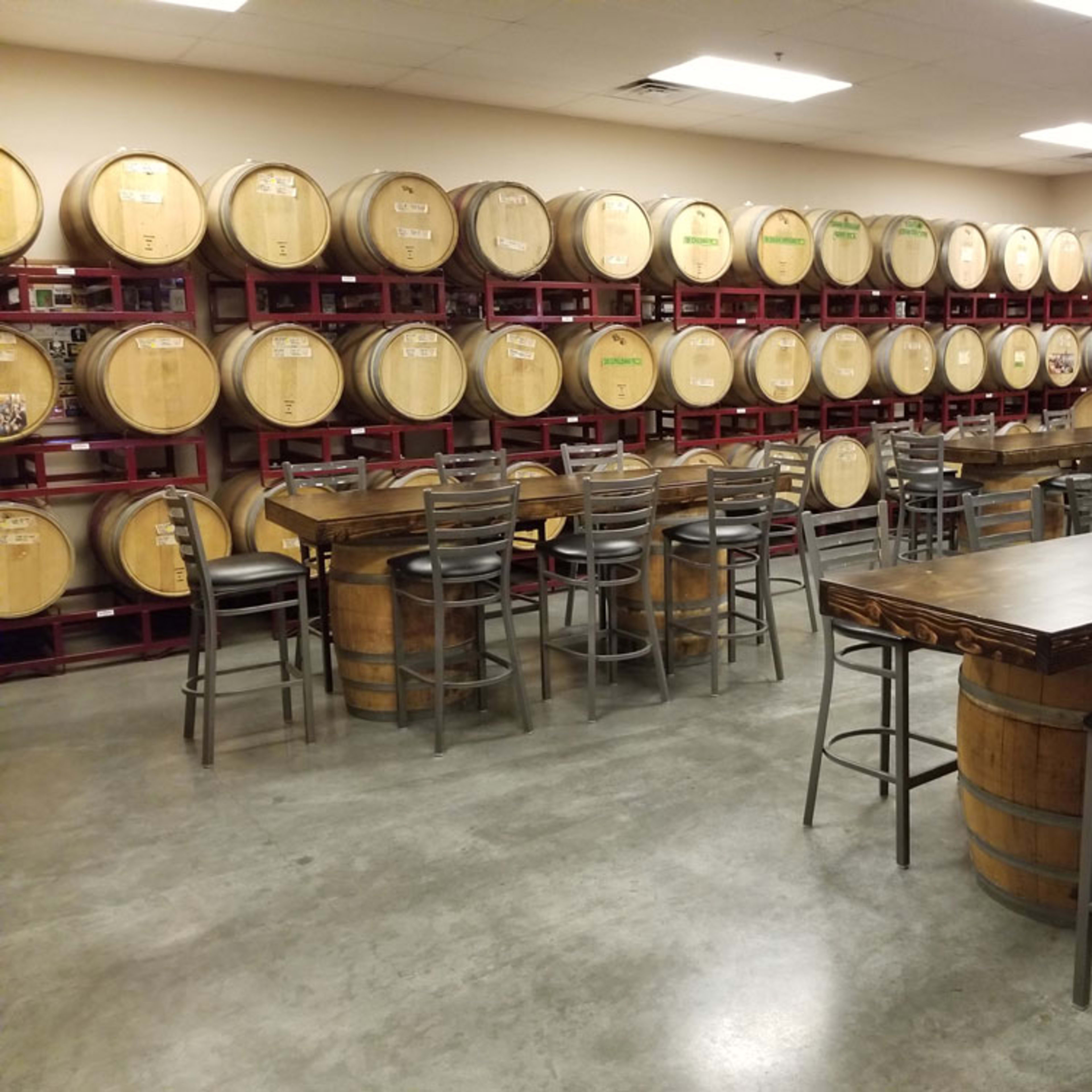 A wine tasting room with wooden barrels along the wall and tables with high stools arranged in the center.