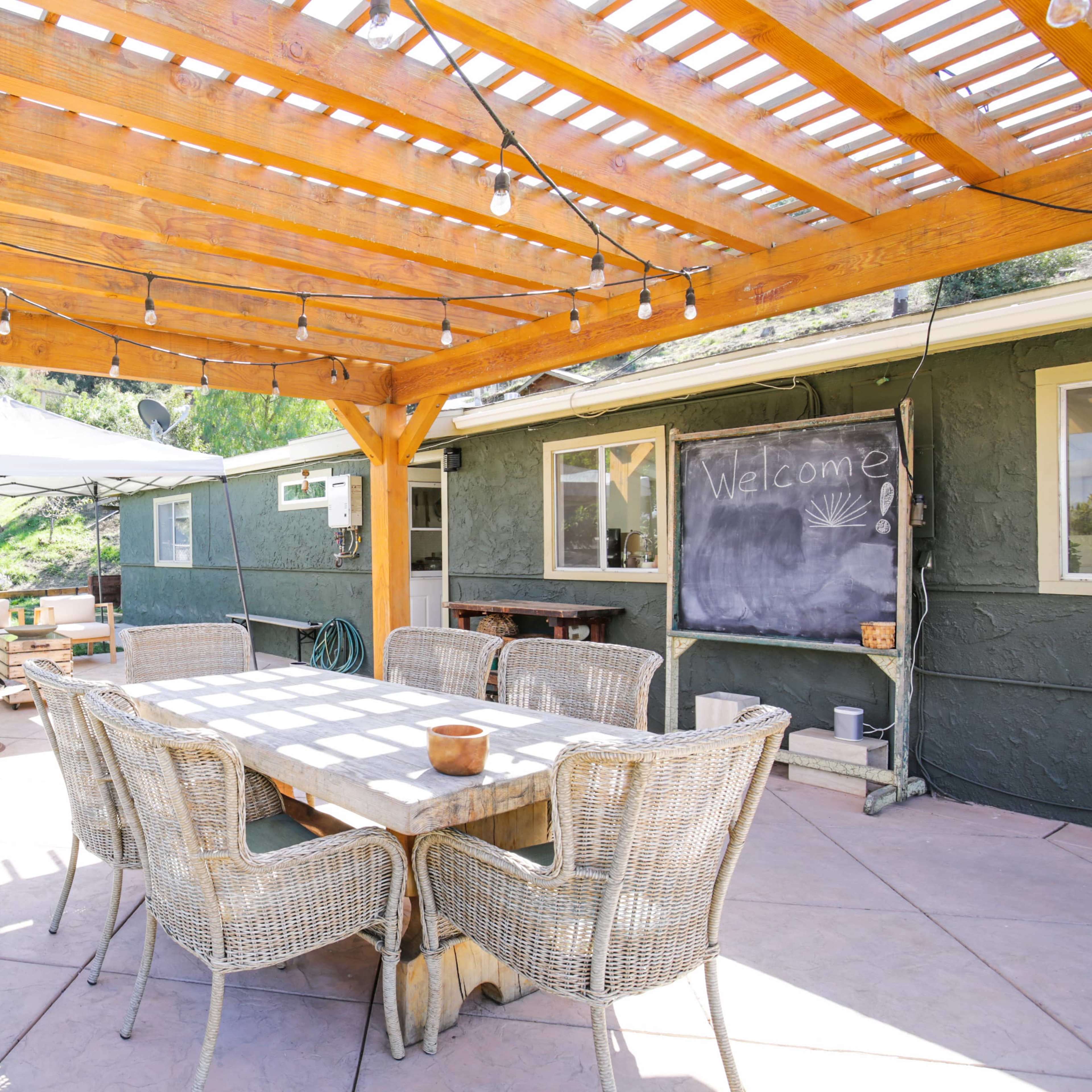 A patio area with a wooden pergola, dining table, and a grill, adjacent to a house surrounded by greenery.