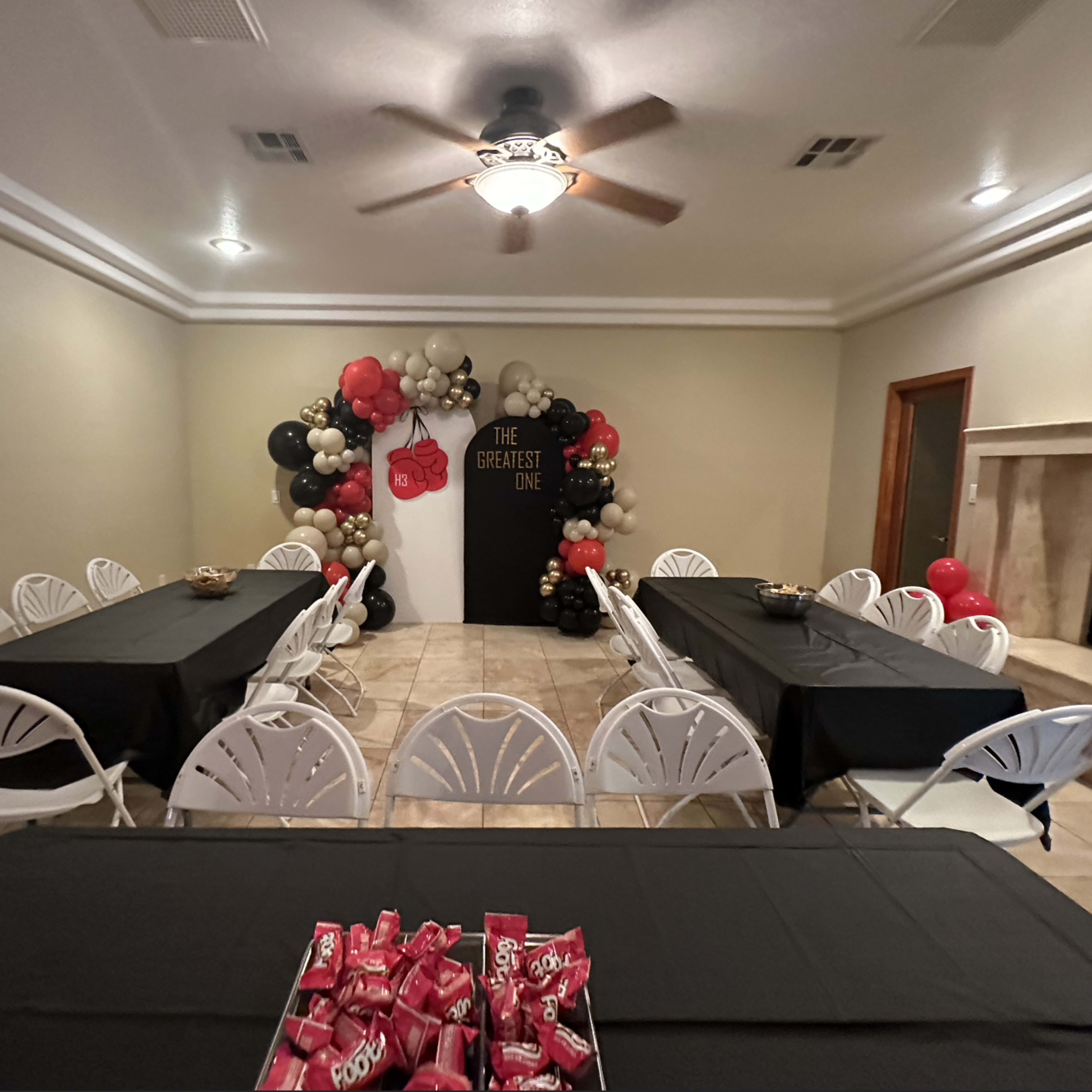 A room is set up for a celebration with multiple black tables and a decorated wall featuring red, black, and gold balloons.