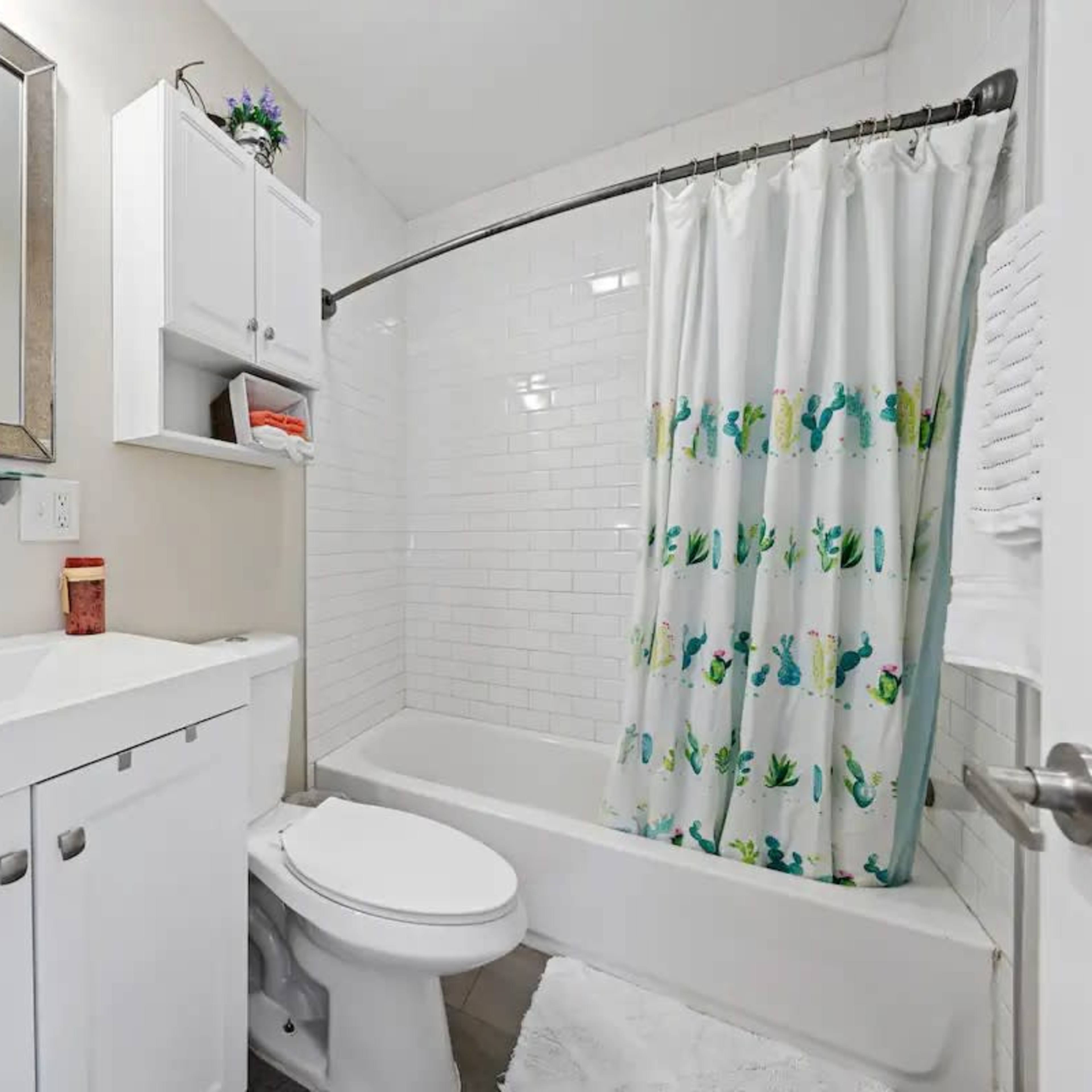 A small bathroom featuring a white vanity with a sink, a toilet, a shower with a curtain decorated with cactus designs, and white tiled walls.