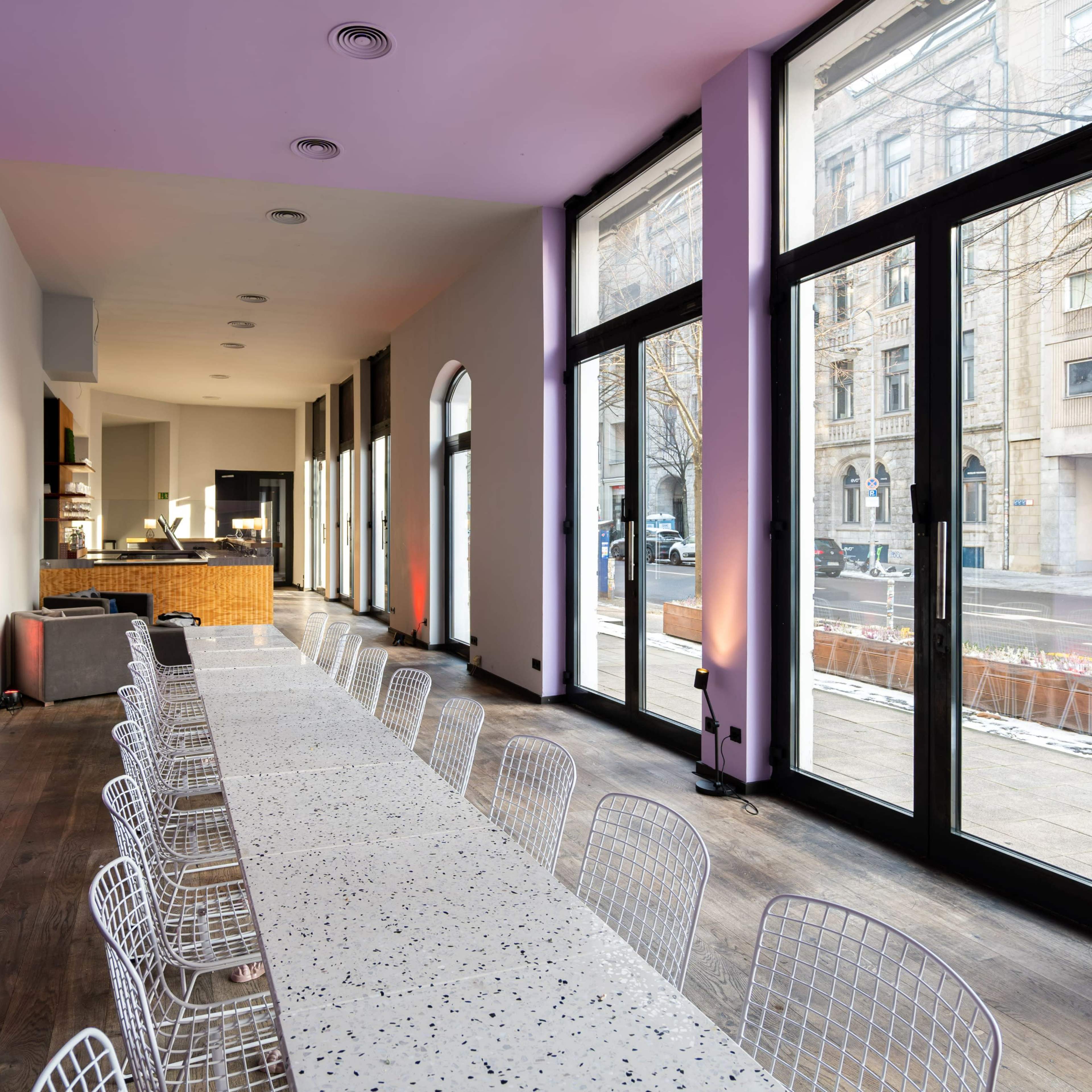 A long table with white chairs is set in a brightly lit room featuring large windows facing an urban street.