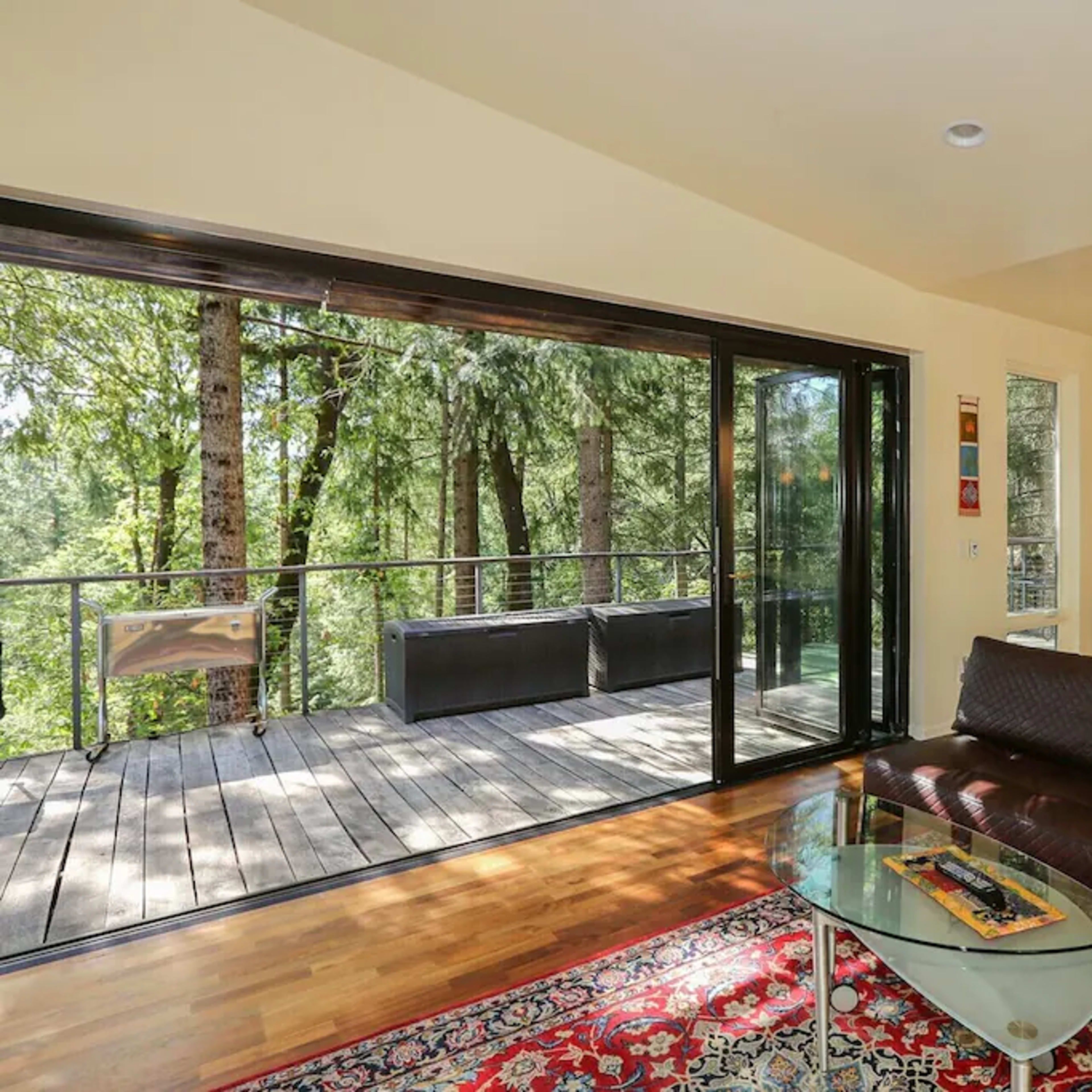 A modern living room with a large glass door opening to a wooden deck surrounded by trees.