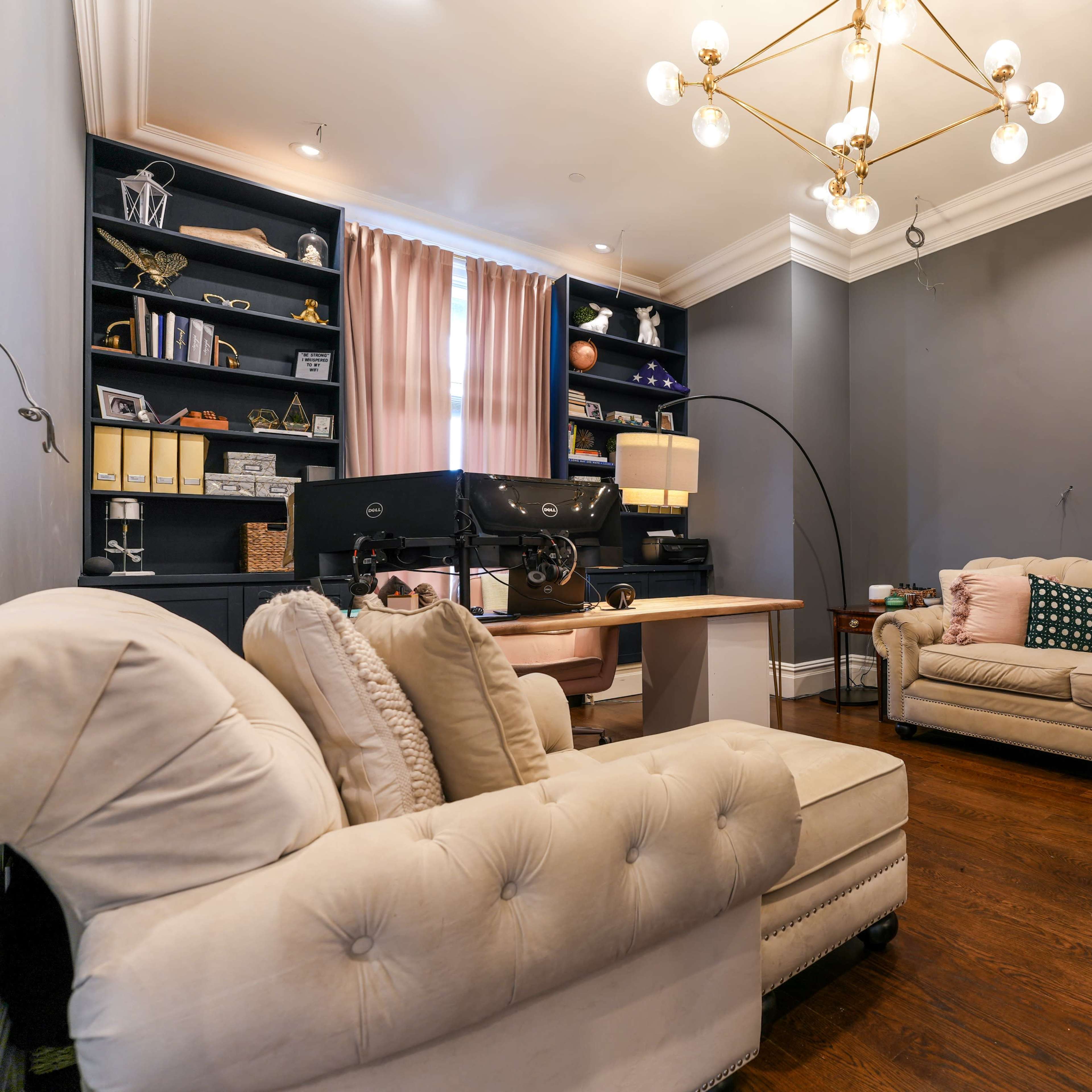 A chic living room featuring two cream-colored sofas, a wooden desk, a large chandelier, and a bookshelf against a gray wall.