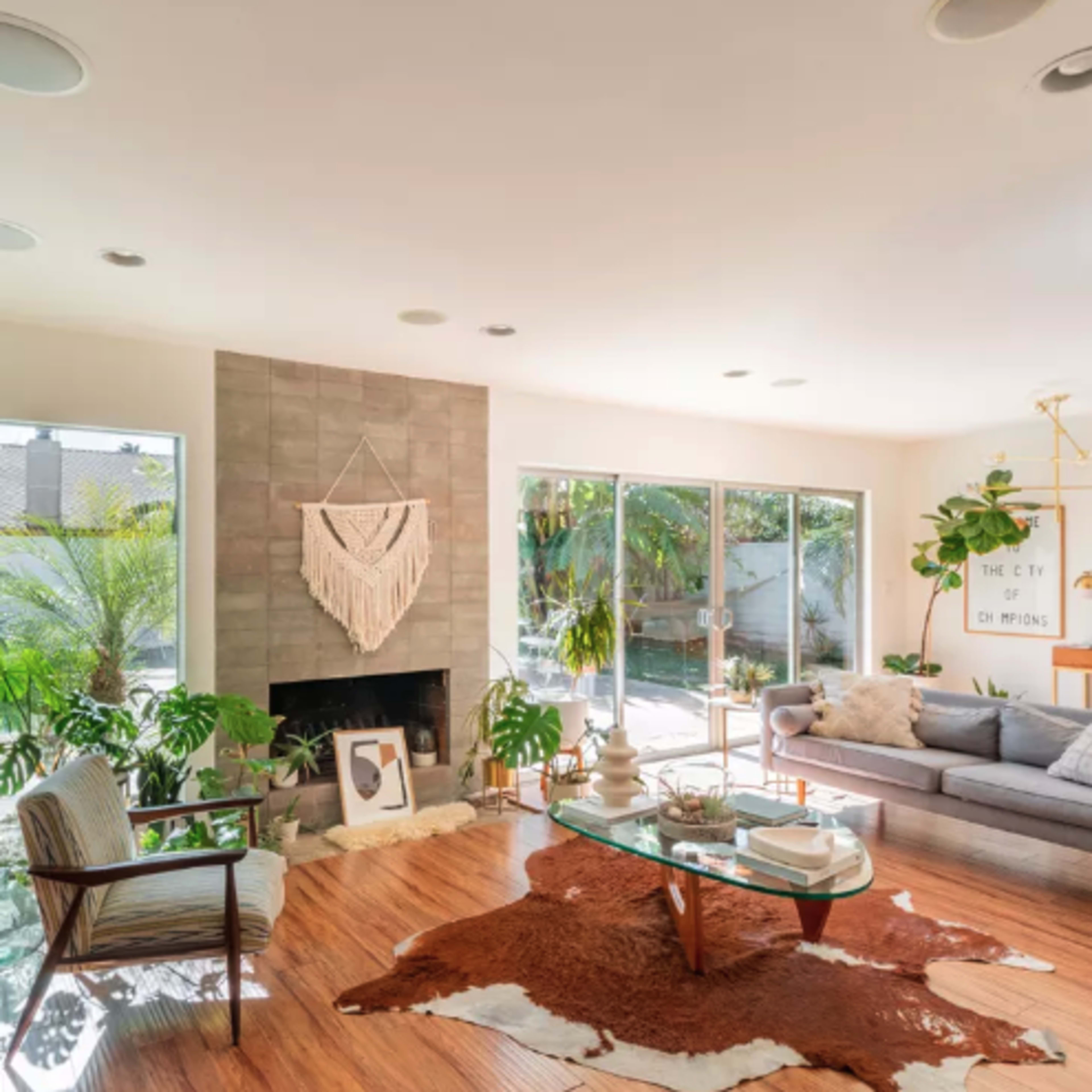 The living room features a grey sectional sofa, a round glass coffee table, and large windows that overlook a patio with greenery.