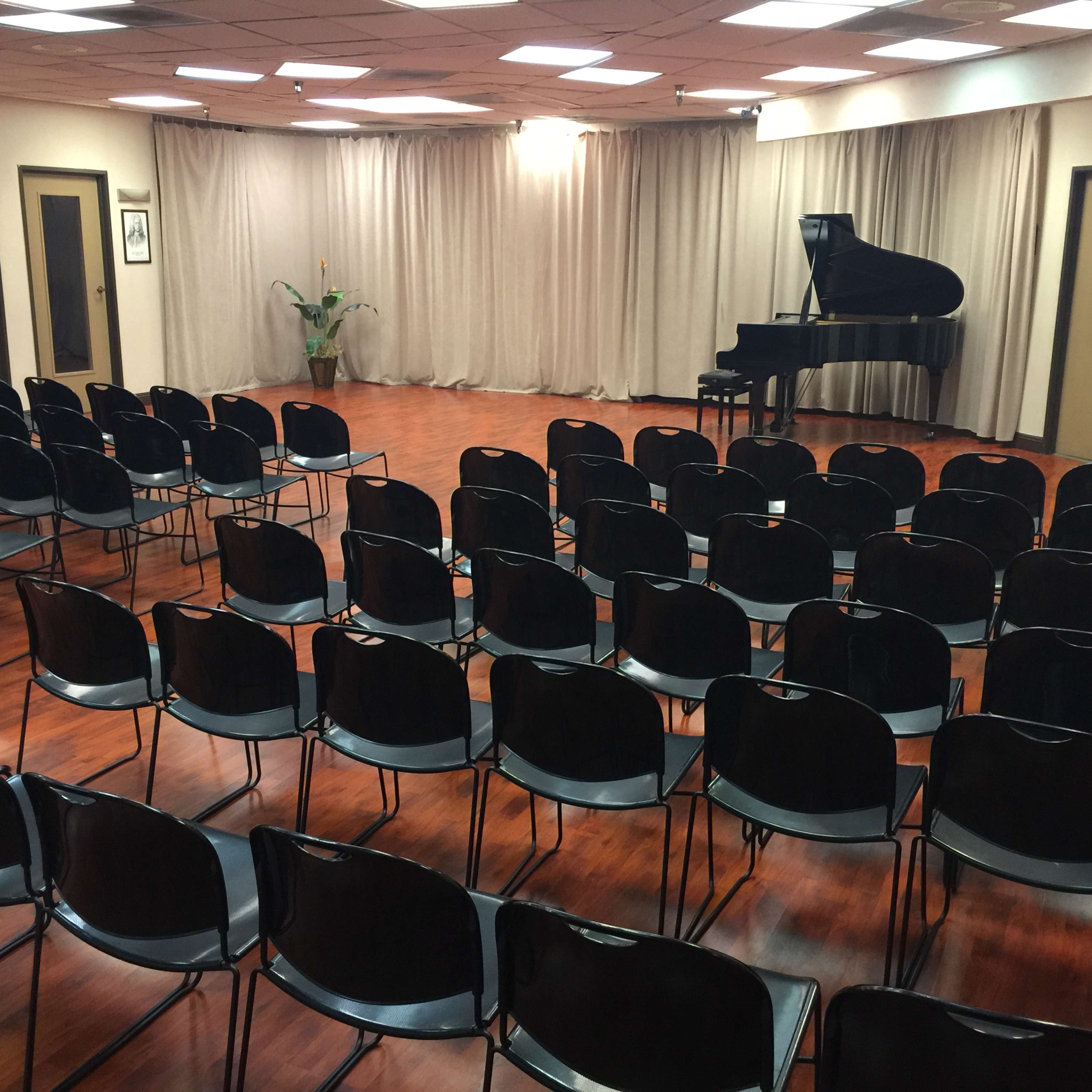 A concert hall setup with rows of black chairs facing a grand piano in front of a curtain.