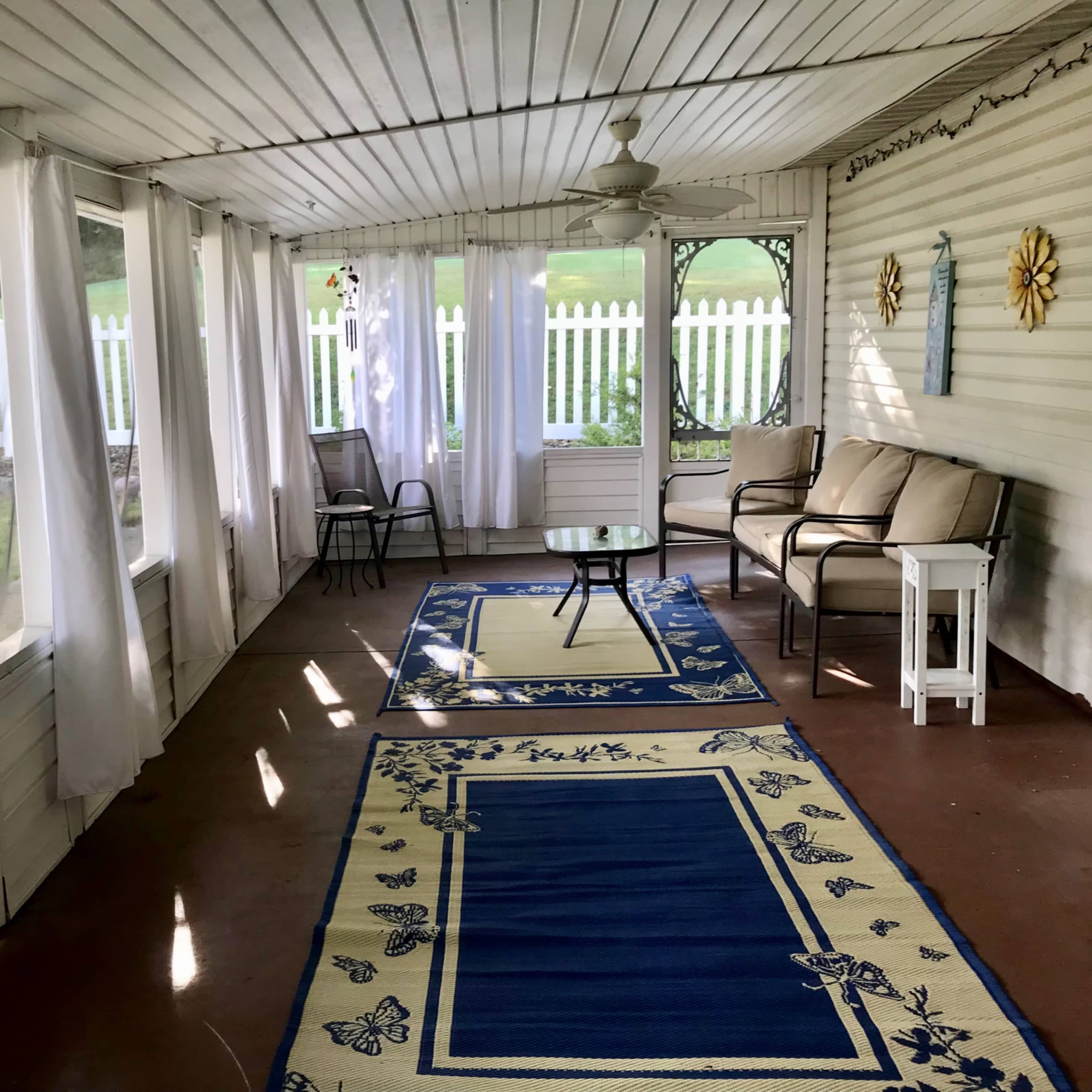 A screened-in porch with two seating areas, a small table, and decorative rugs against a backdrop of white picket fencing and greenery outside.