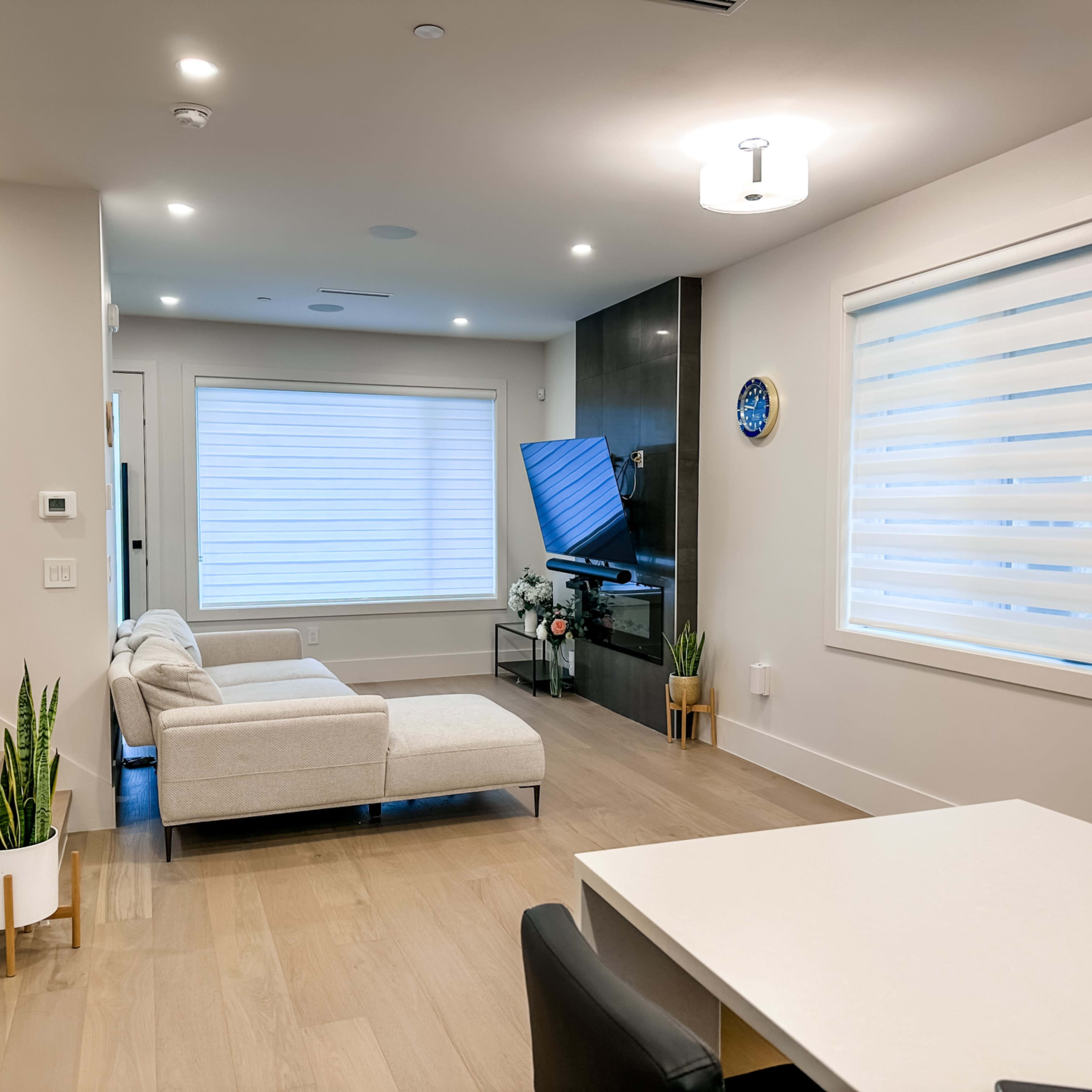 A modern living room featuring a light-colored sectional sofa, a television on a dark wall, and a dining area with a white table and black chairs.