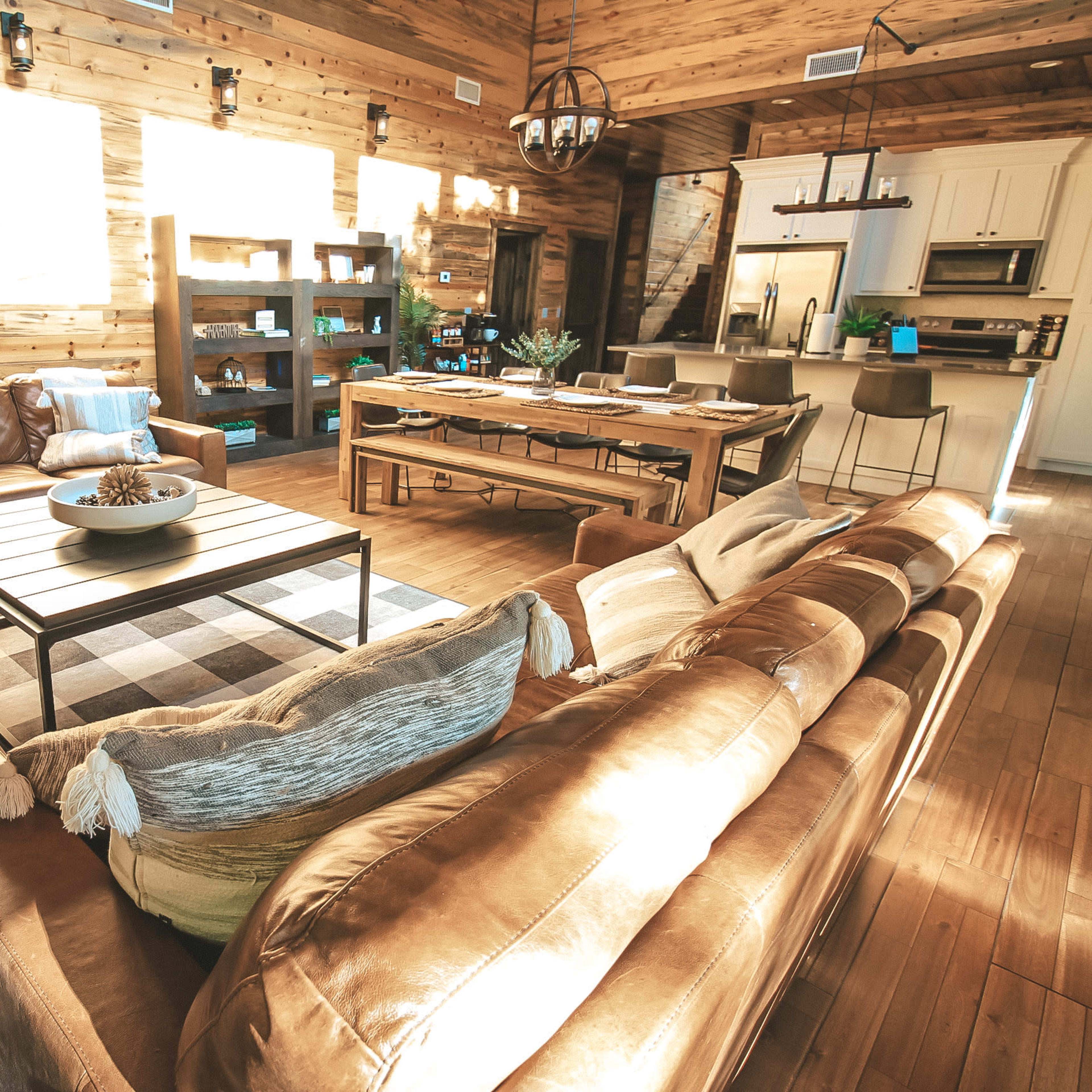 A warm, rustic living room with leather sofas, a wooden dining table, and large windows that let in natural light.