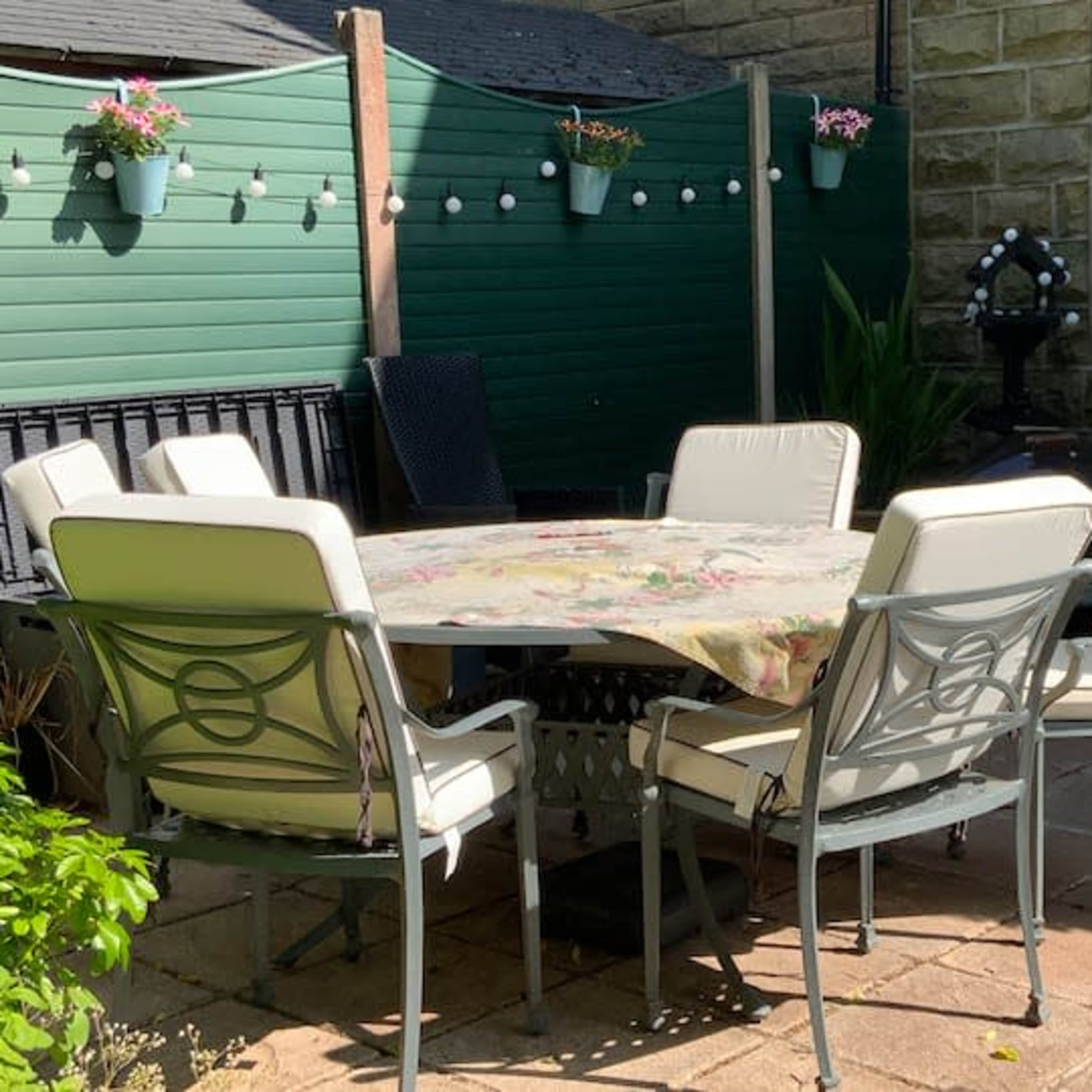 A round table surrounded by six metal chairs is set in a patio area, with a green fence and potted plants in the background.