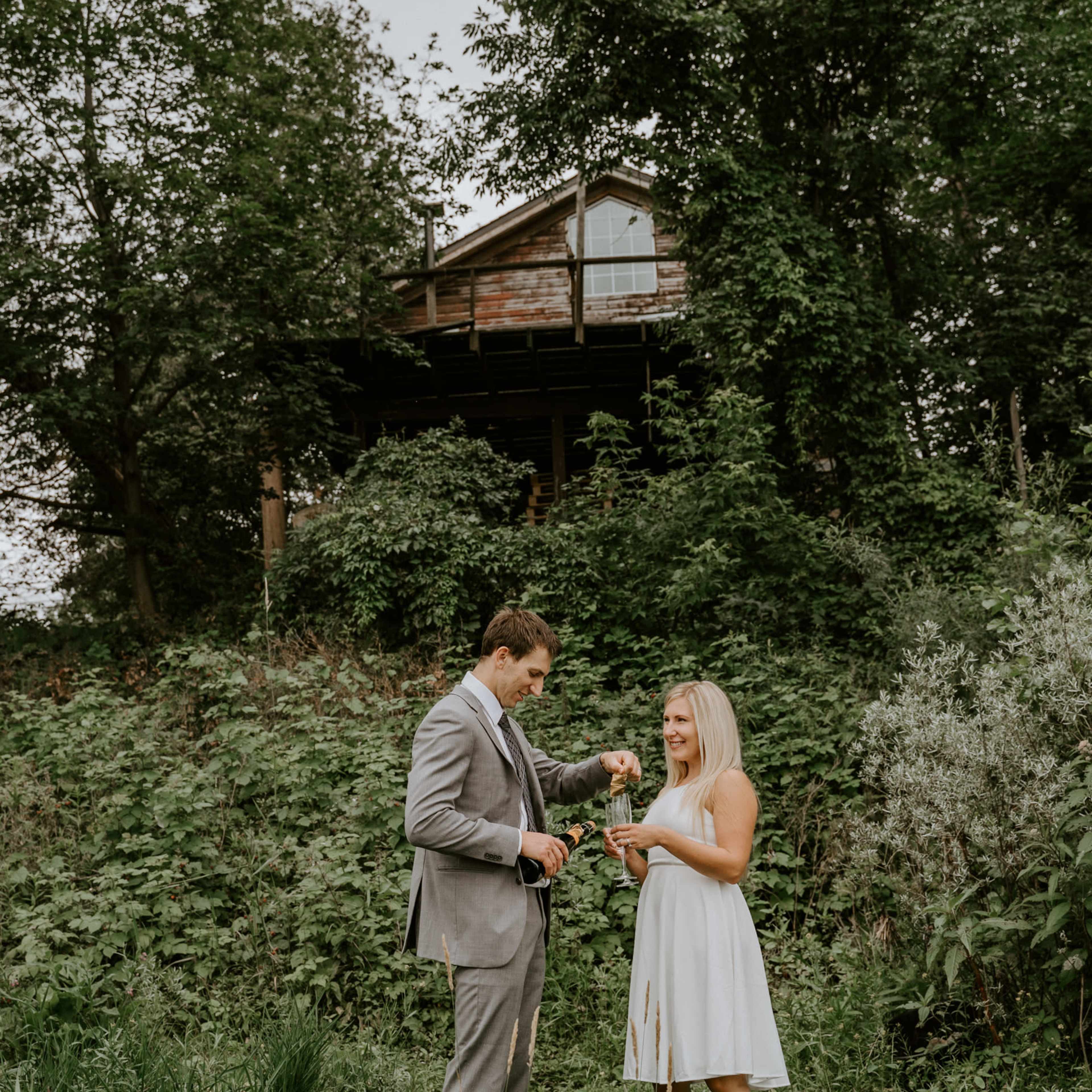 A man in a suit and a woman in a white dress stand on a grassy area near a house surrounded by trees.