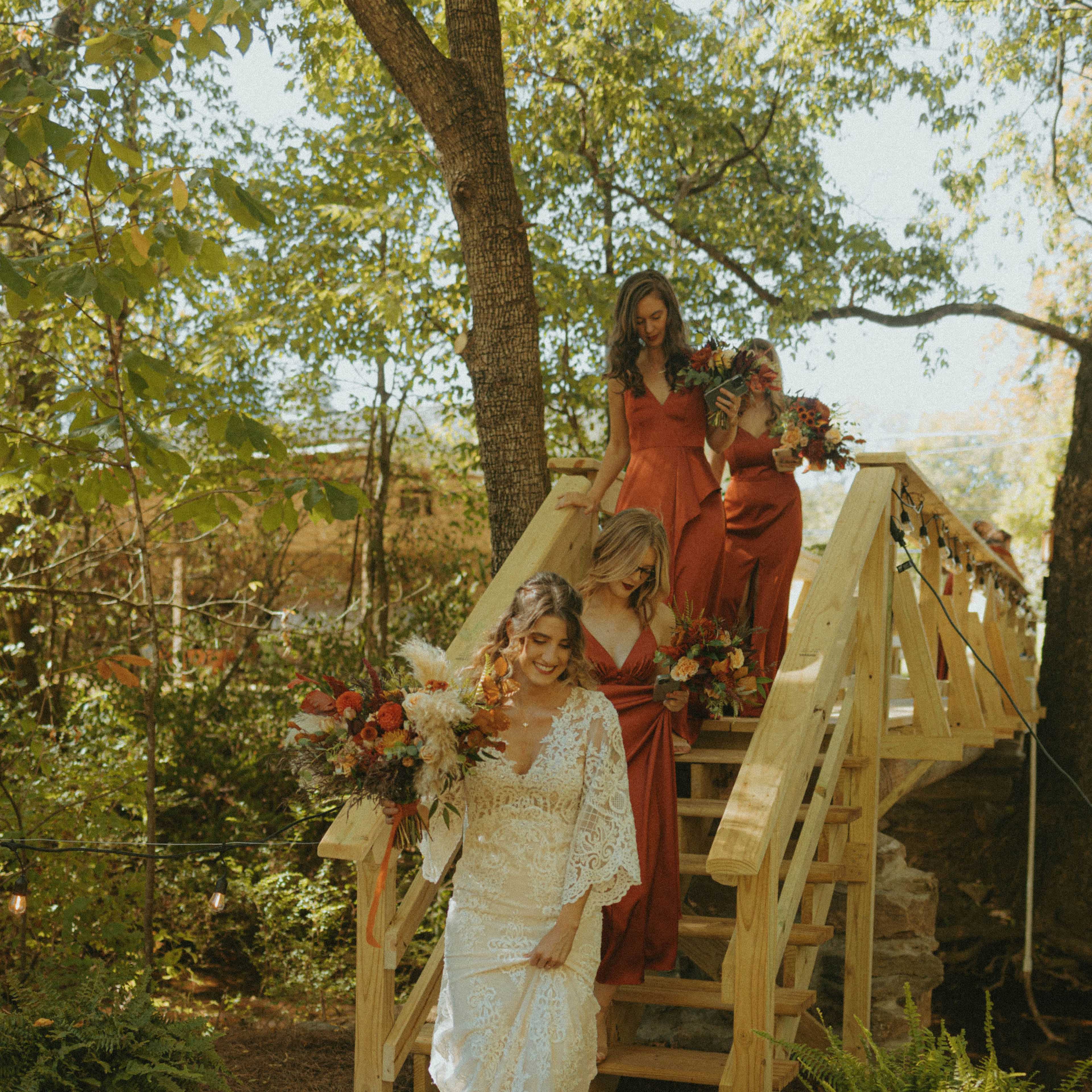A bride in a white lace dress descends wooden stairs accompanied by three women in red dresses, each holding floral arrangements, surrounded by greenery.