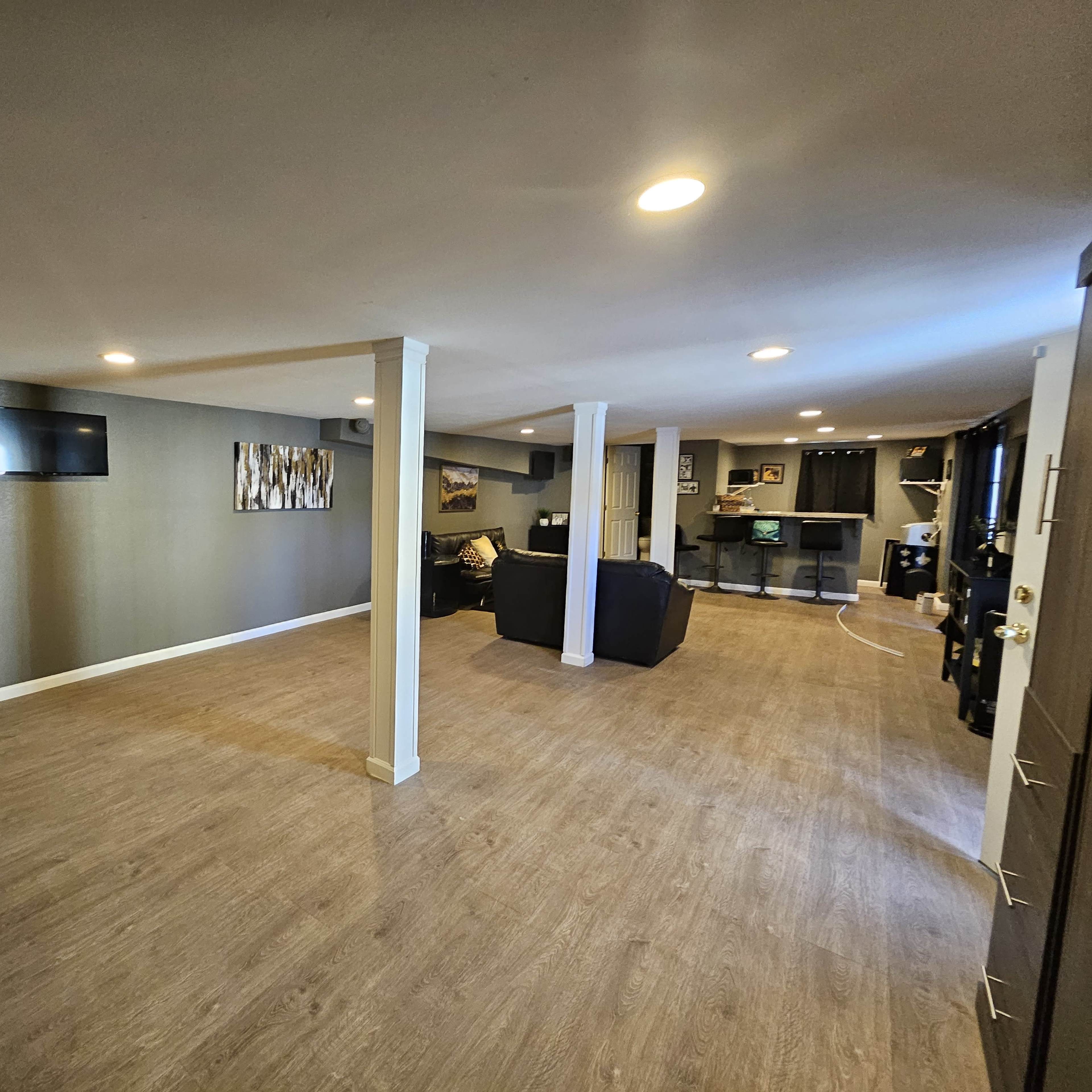 A spacious basement with wooden flooring, a small bar area in the background, and a wall-mounted television.