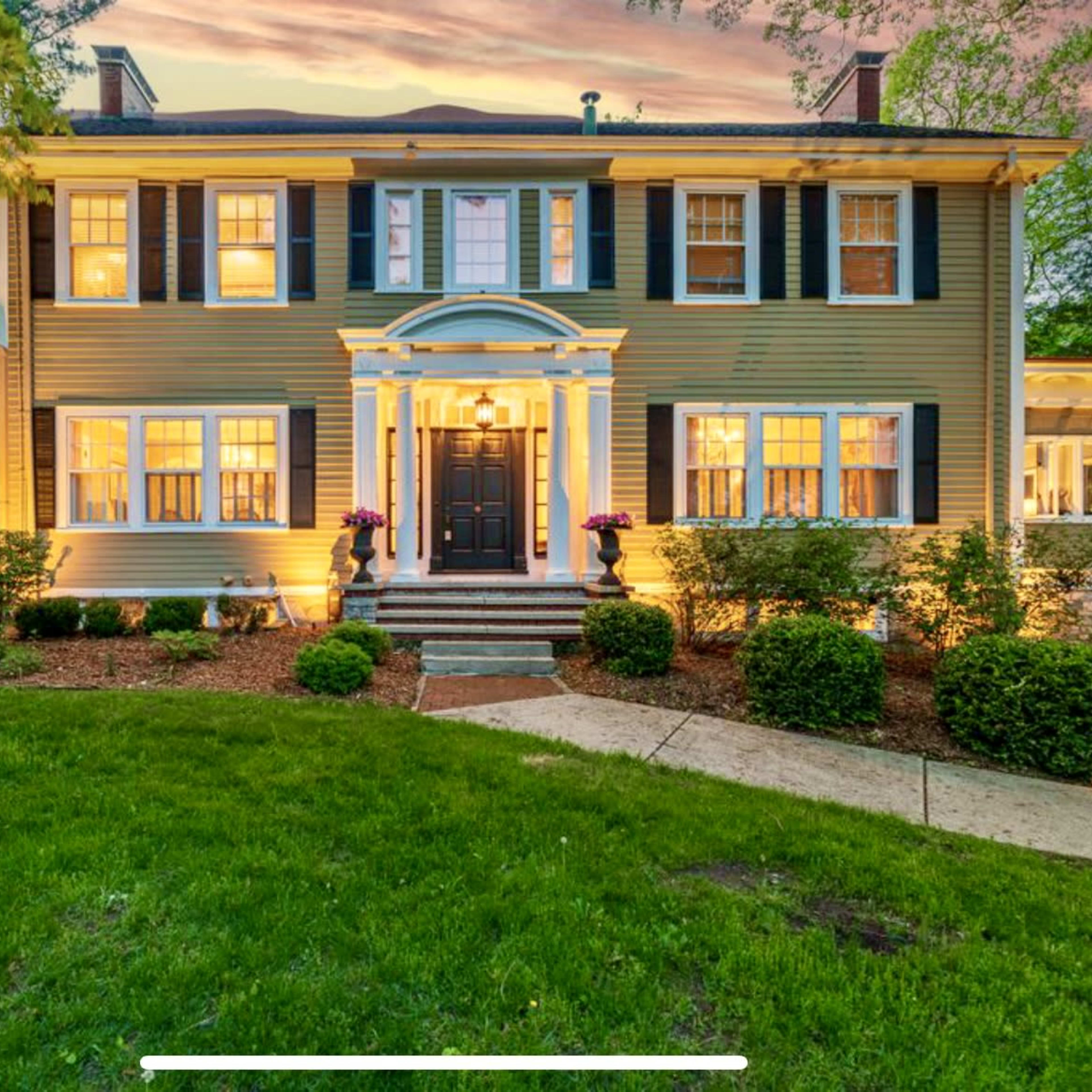 A two-story house with light green siding, white trim, and well-landscaped gardens illuminated by evening light.