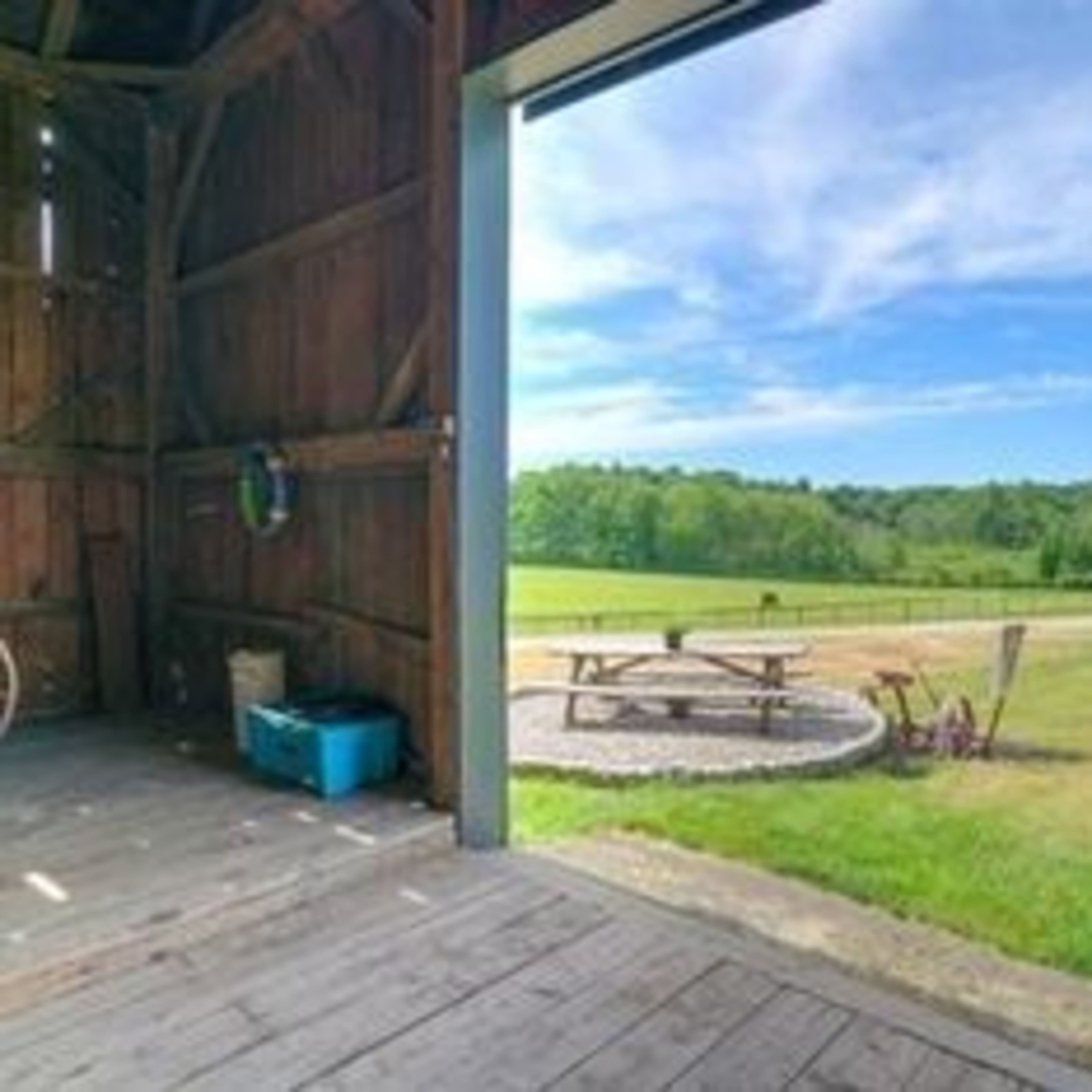 The interior of a barn with an open door that reveals a grassy field and a picnic table surrounded by a circular stone pathway.