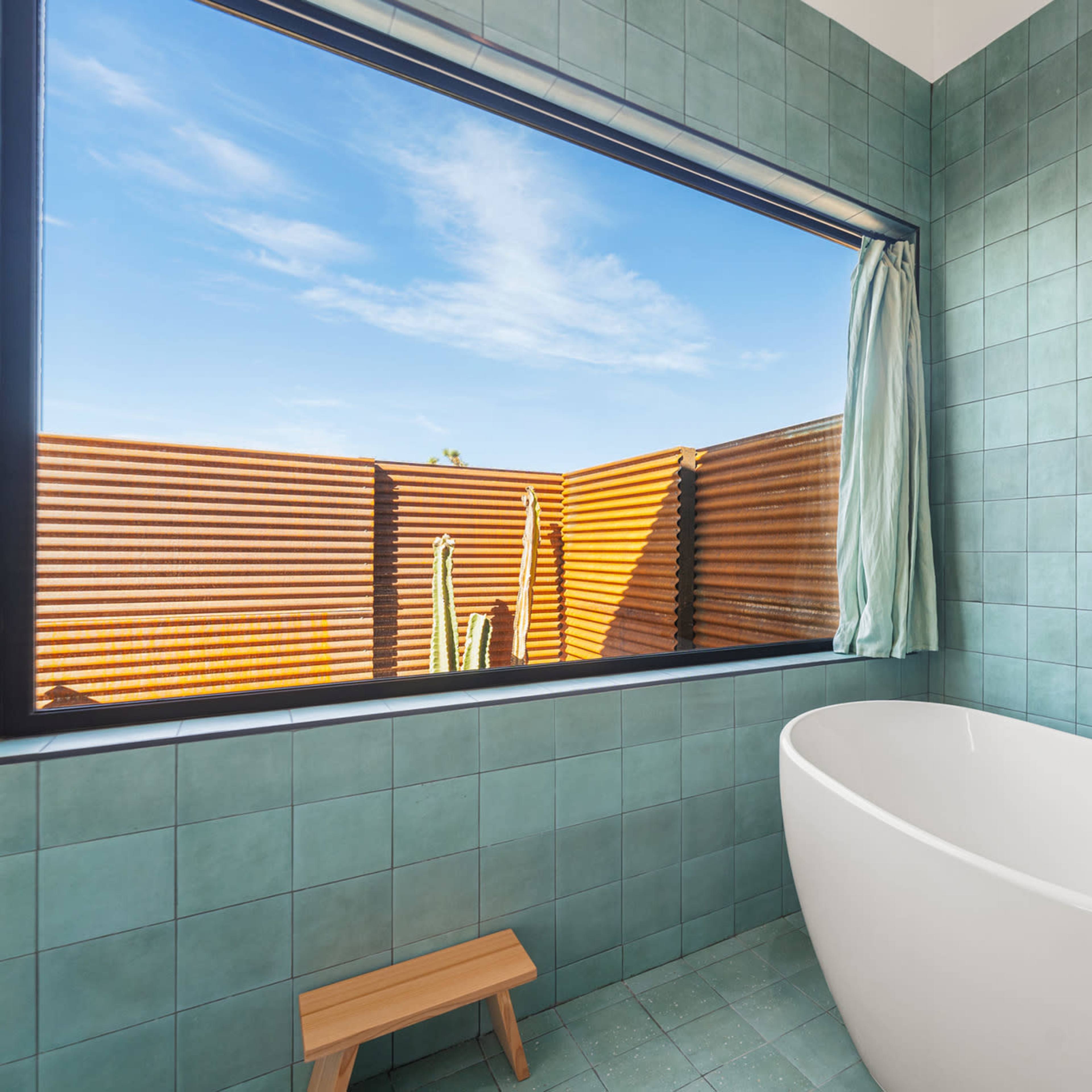 A modern bathroom features a freestanding white bathtub beside a large window with a view of a wooden fence and cacti.