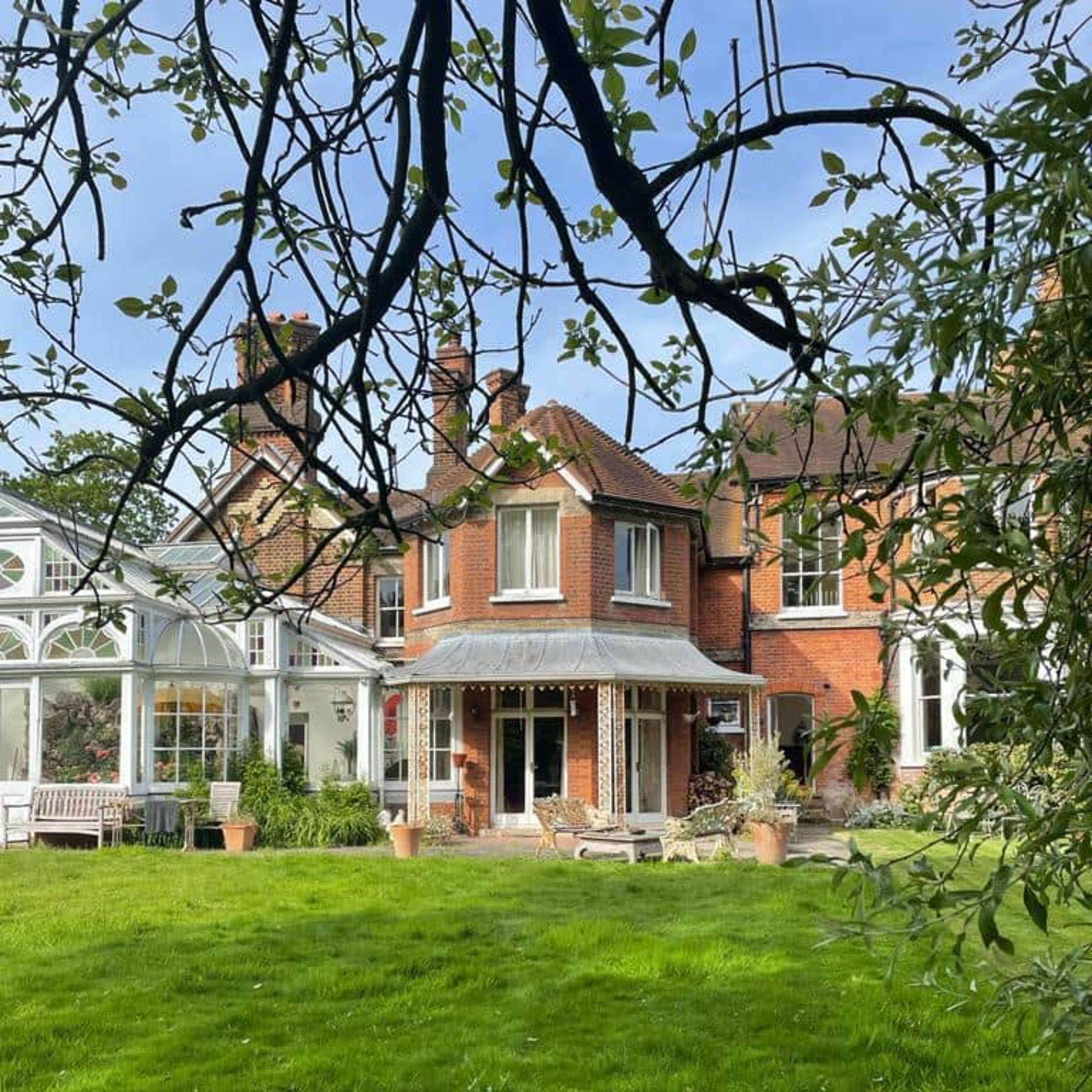 A brick house with multiple sections, including a circular turret, is set in a green lawn surrounded by trees and plants.