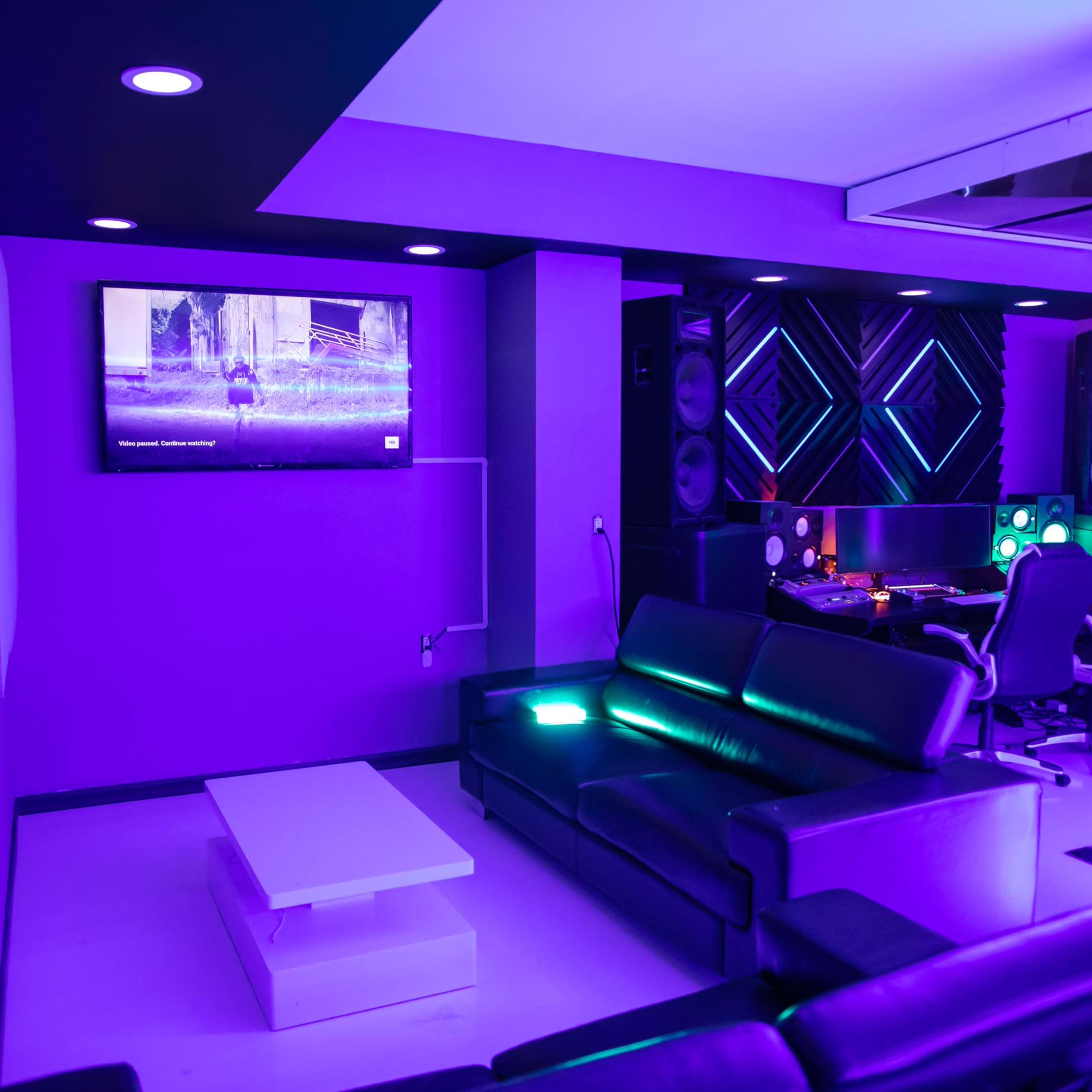 A modern media room with purple lighting, a black sofa, a coffee table, and a workspace featuring audio equipment and a television on the wall.