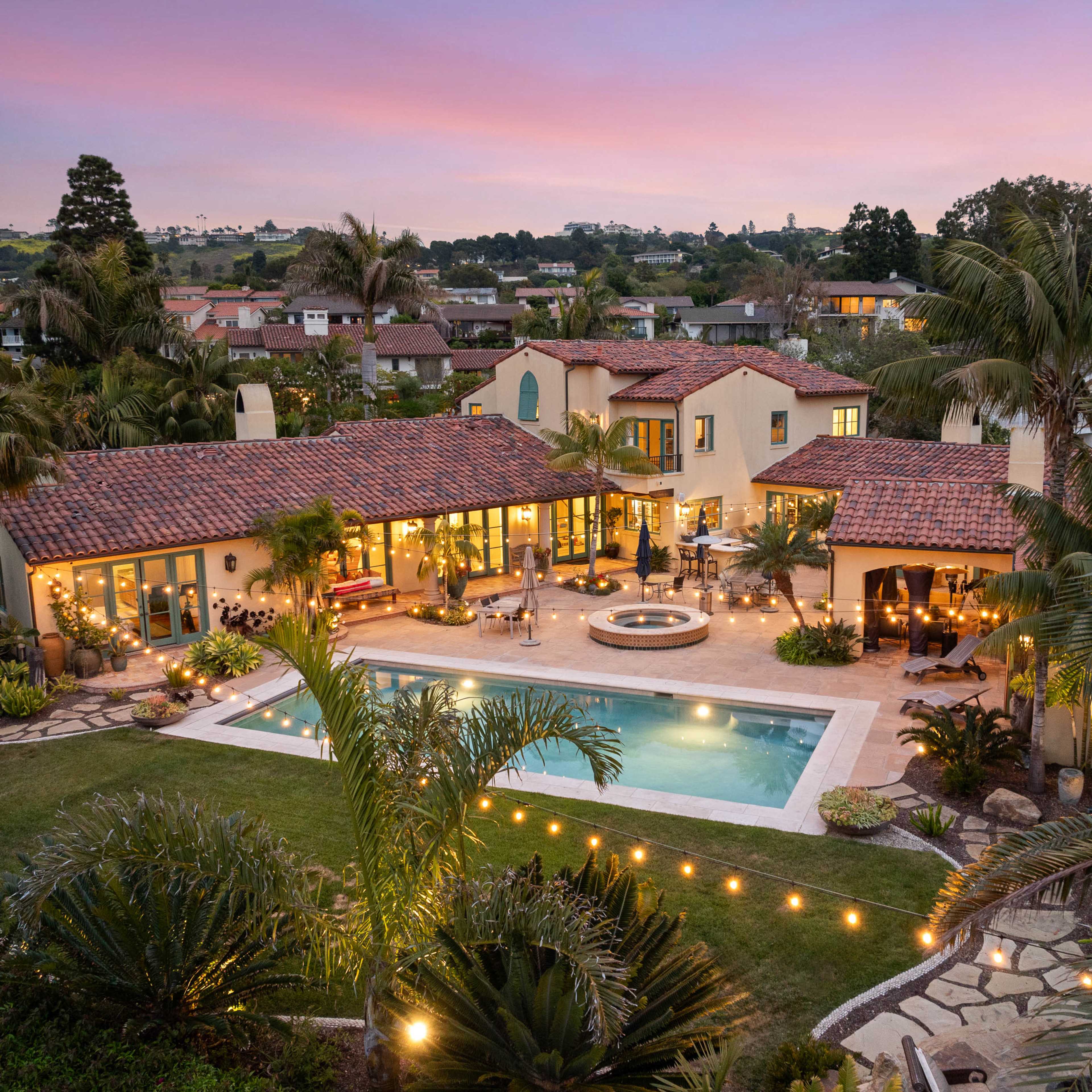 A luxurious home with a tiled roof surrounds a large pool and landscaped garden, set against a vibrant sunset sky.