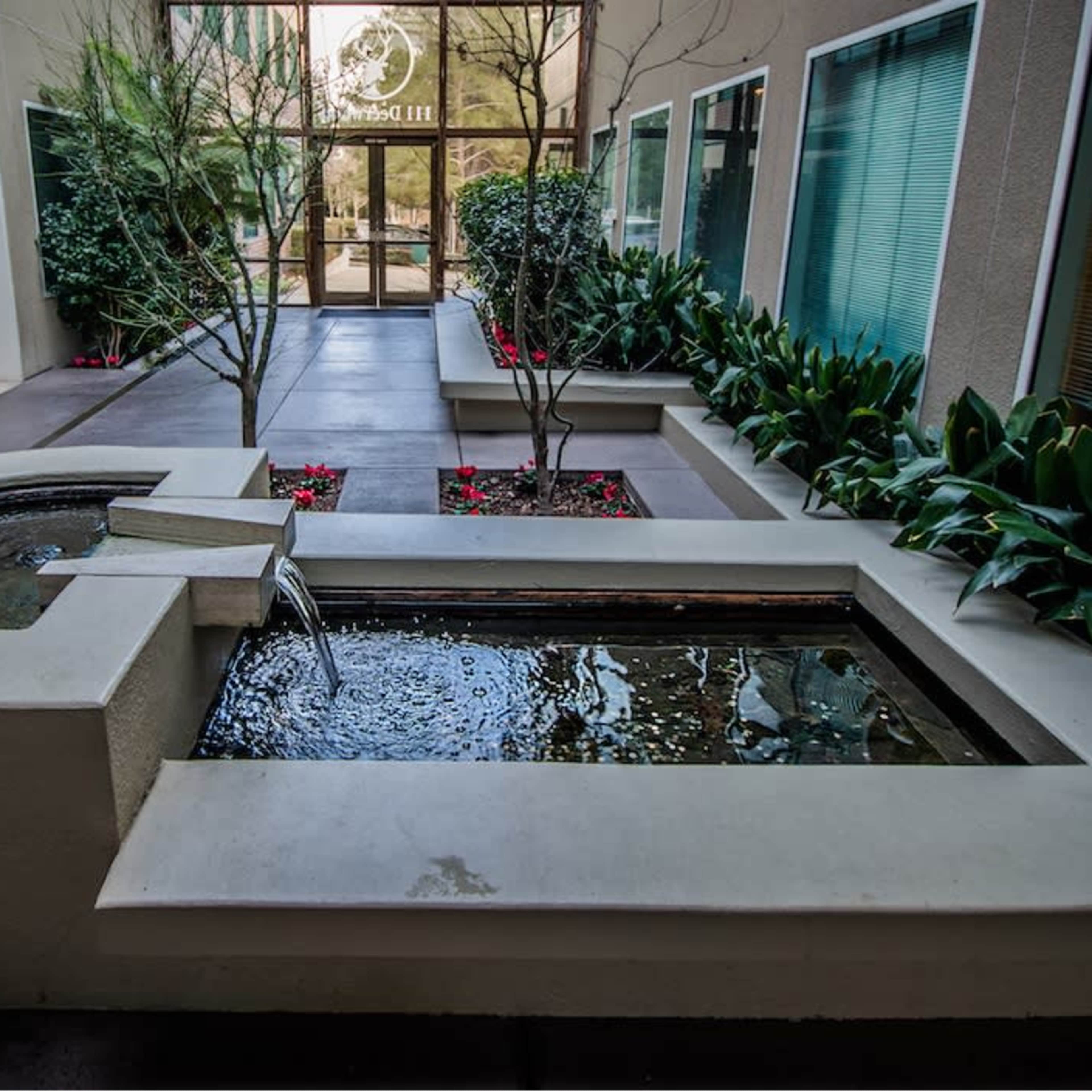 A courtyard with a water feature and greenery, leading to a glass entrance with a logo.