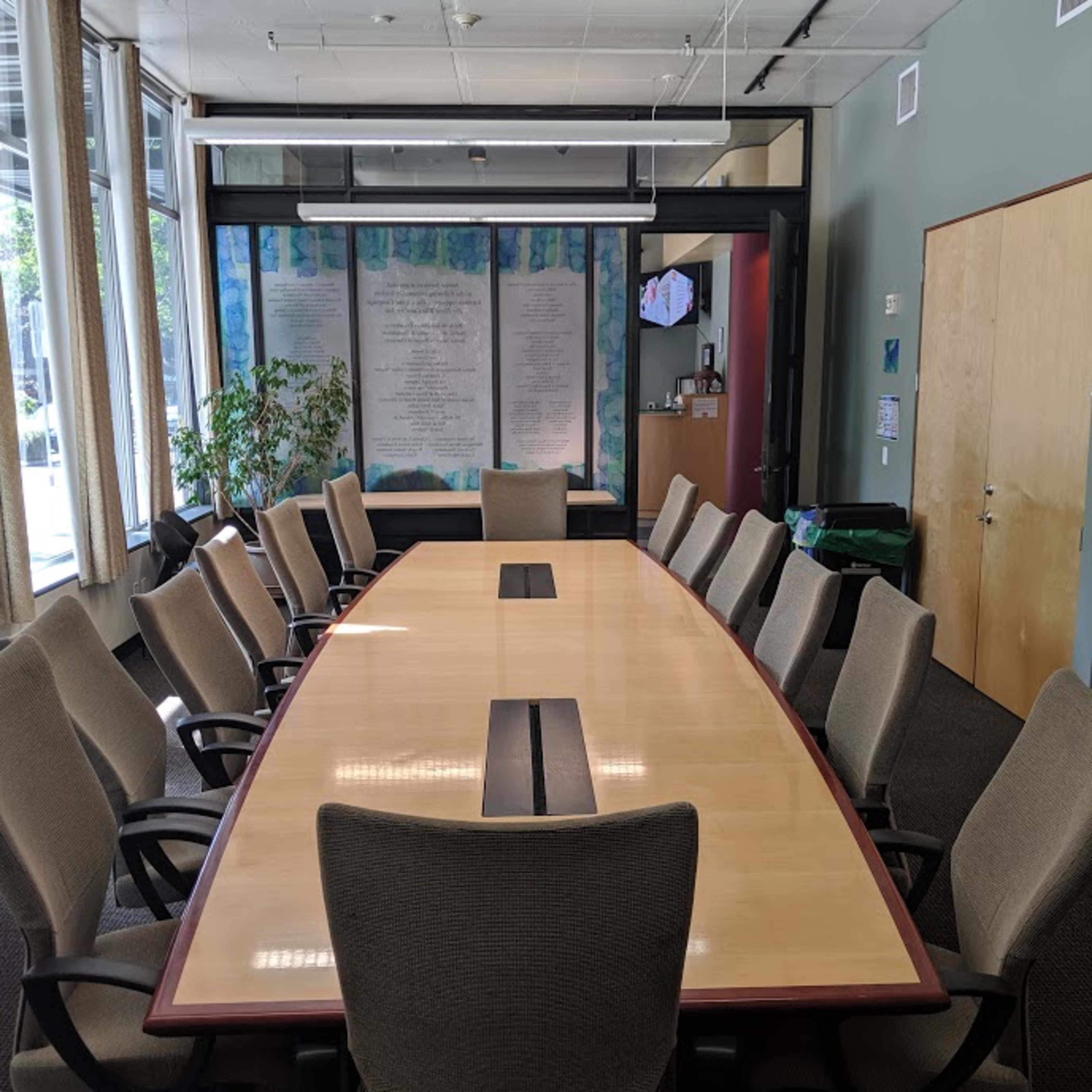A large conference room features a rectangular table surrounded by gray office chairs, with a projector mounted on the ceiling and walls adorned with large windows.
