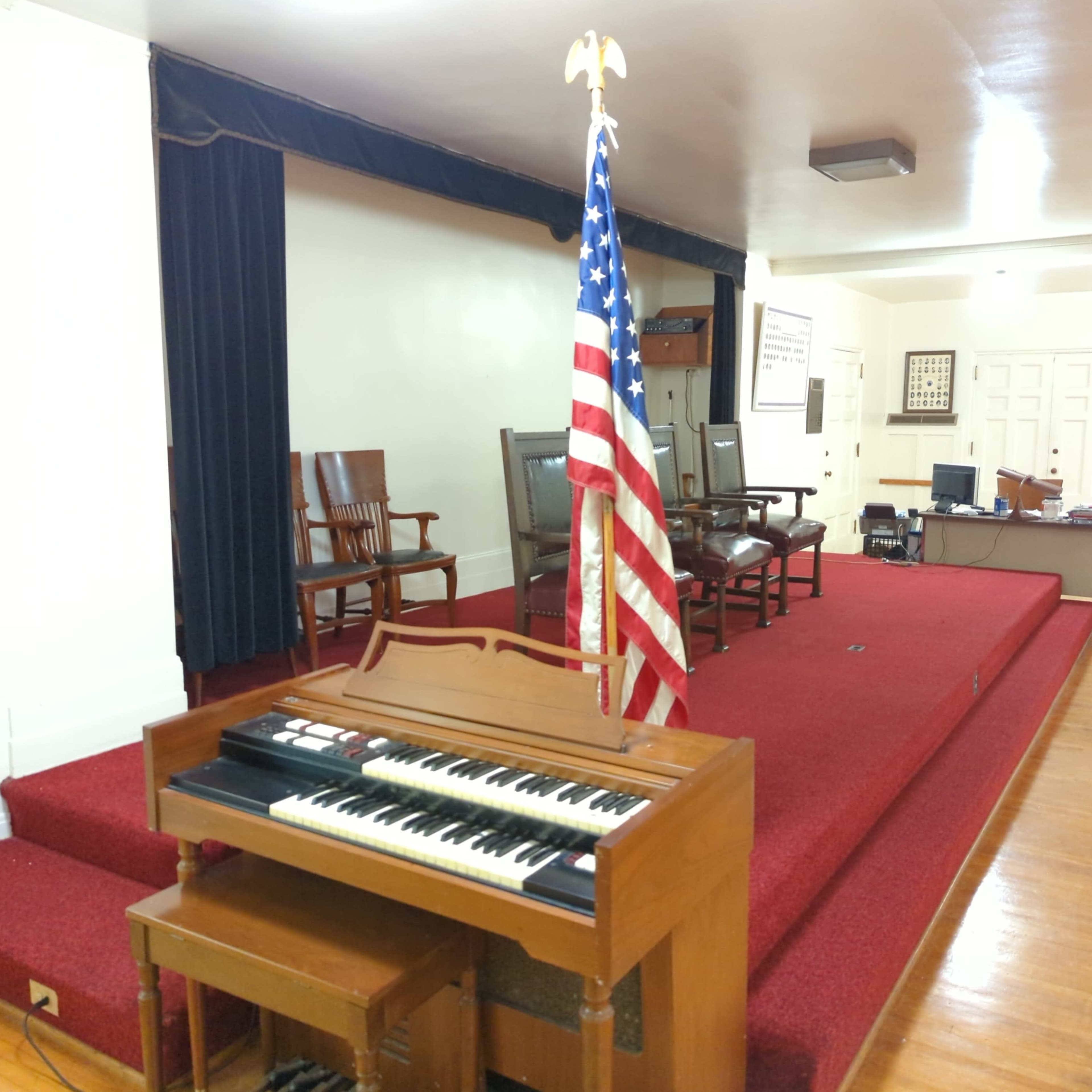 A wooden organ sits in front of a raised platform with several empty chairs and an American flag in a spacious meeting room.