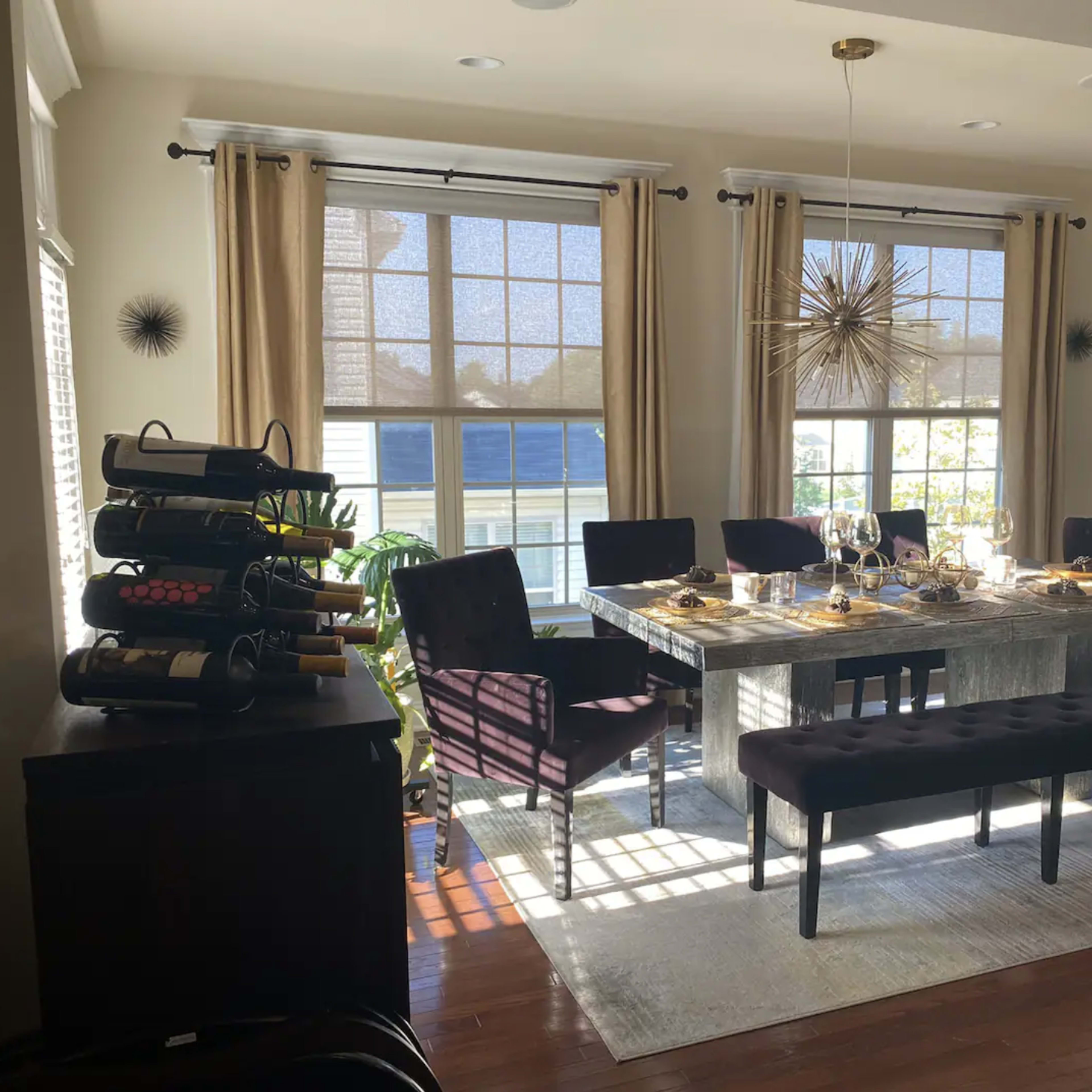 A dining room features a large table set for a meal with wine bottles displayed on a rack and sunlight streaming through large windows.