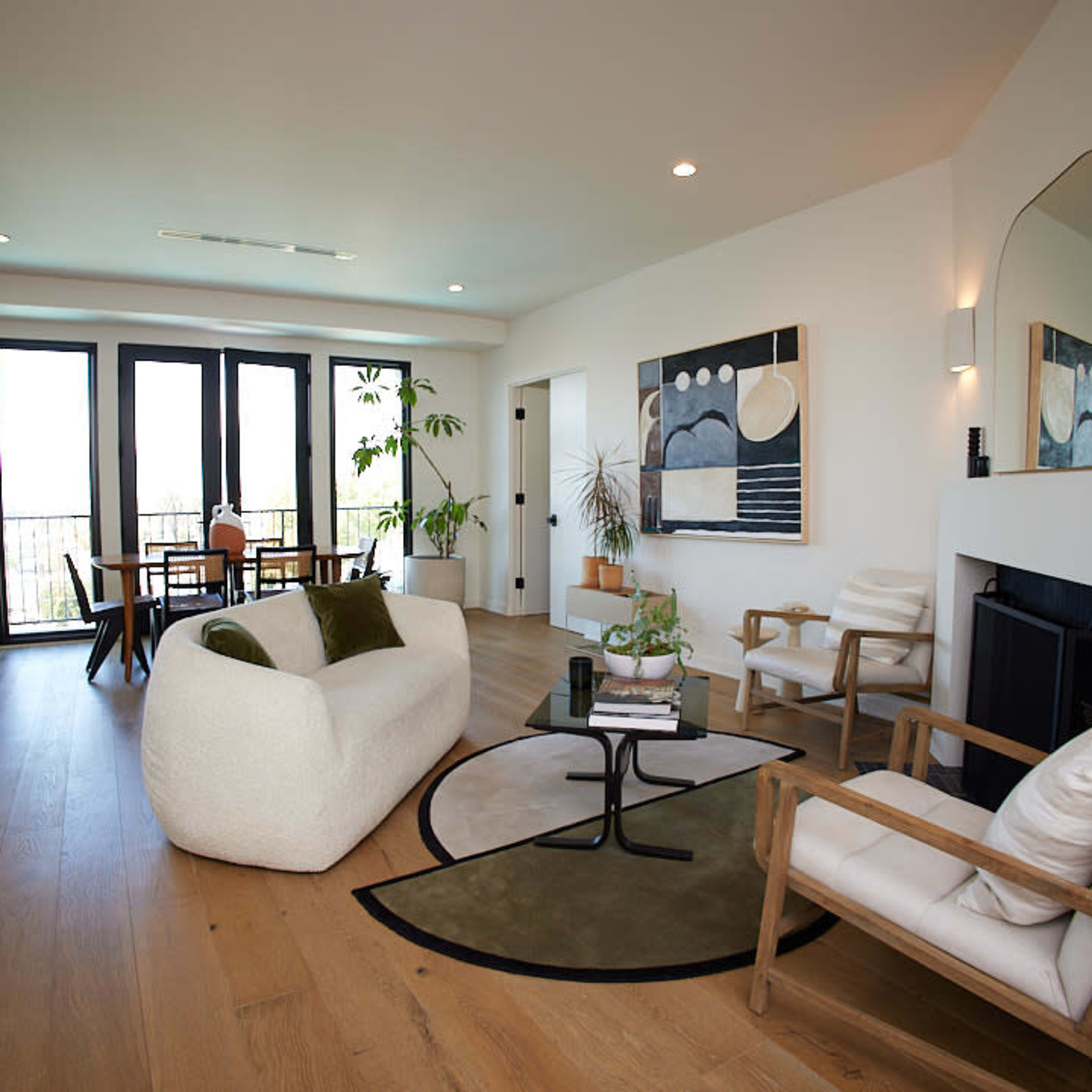 The spacious living room features a cozy white sofa, two armchairs, a round coffee table, and large windows that provide natural light.