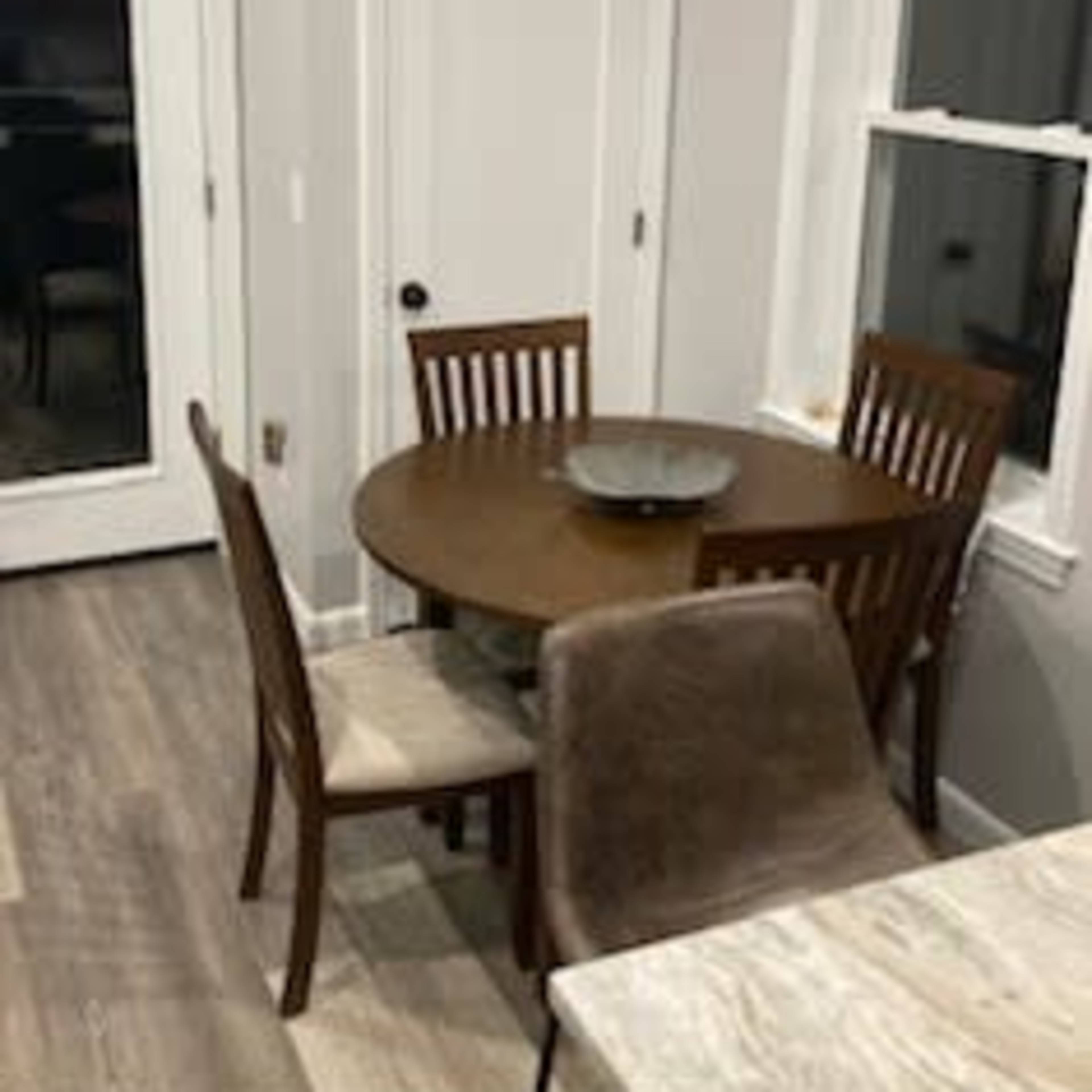 A small wooden dining table with four chairs is positioned in a well-lit corner of a room, next to a door and a window.